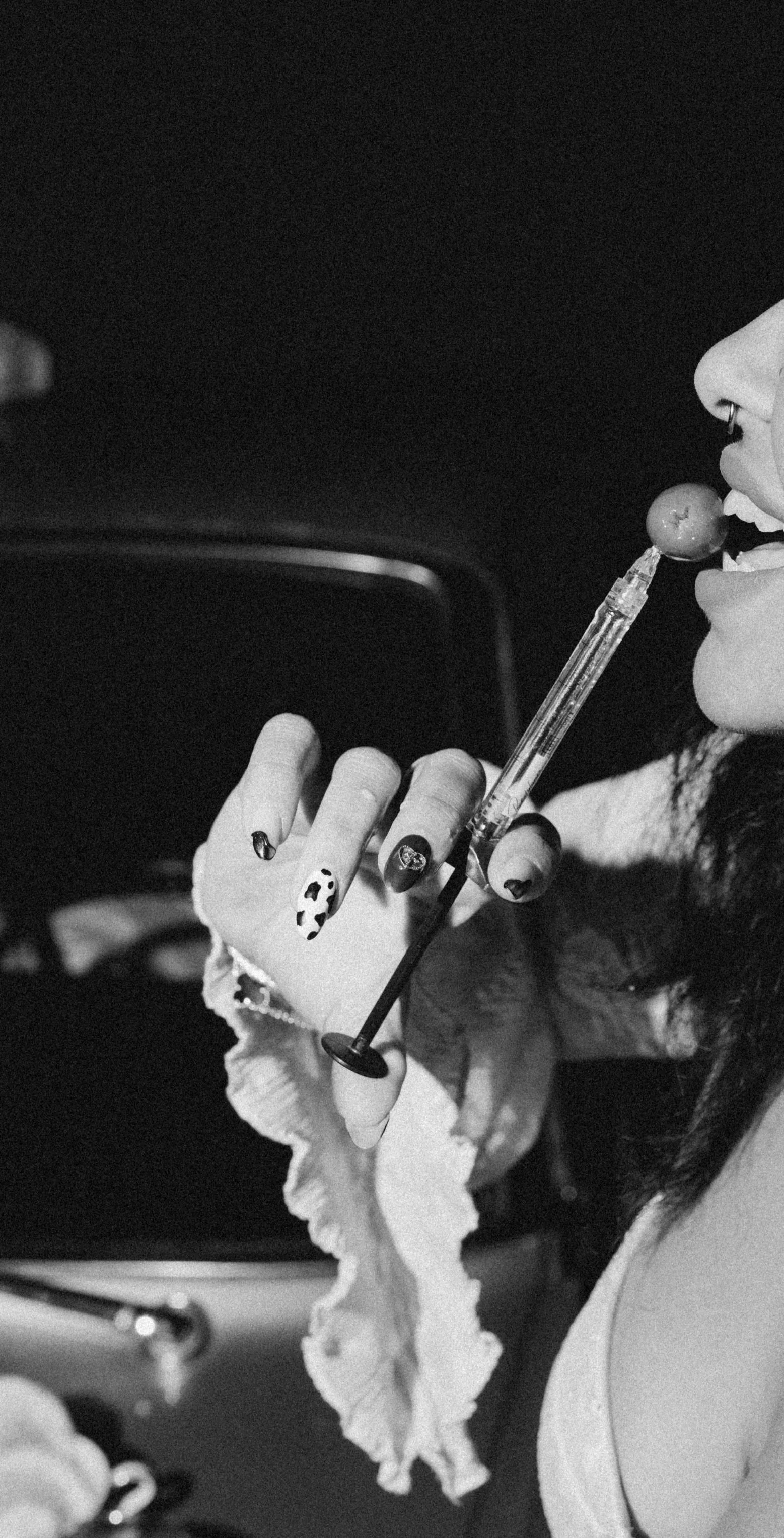 Close-up of a woman with dark hair and nail art, holding and injecting a syringe into her mouth while sitting in a car.