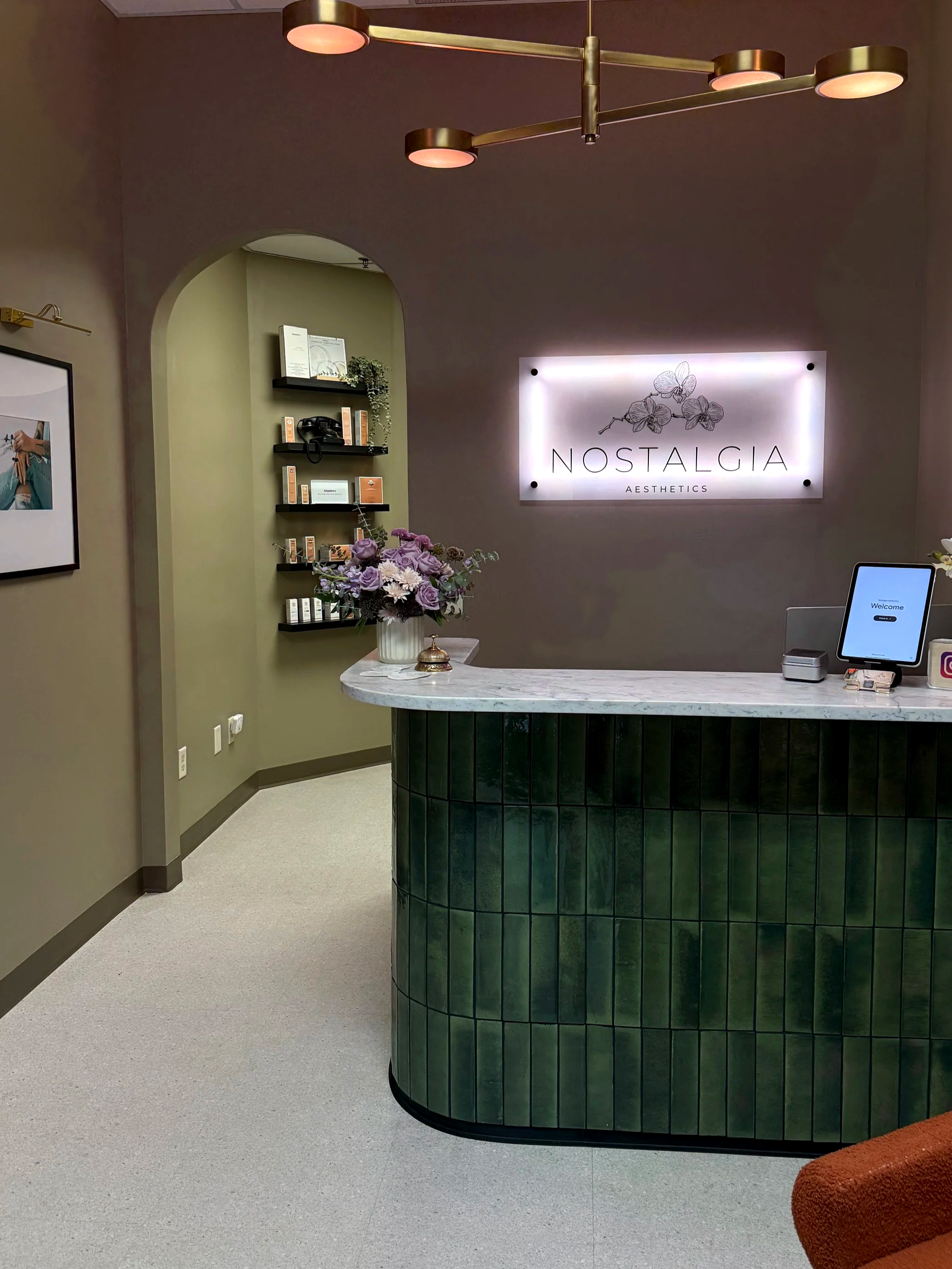 Interior of a reception area with a neon sign 'Nostalgia Aesthetics' on the wall, a curved green tiled front desk with a white marble top, a vase with lavender and white flowers, a tablet on the counter, and a floral wall decor to the left of the sign.