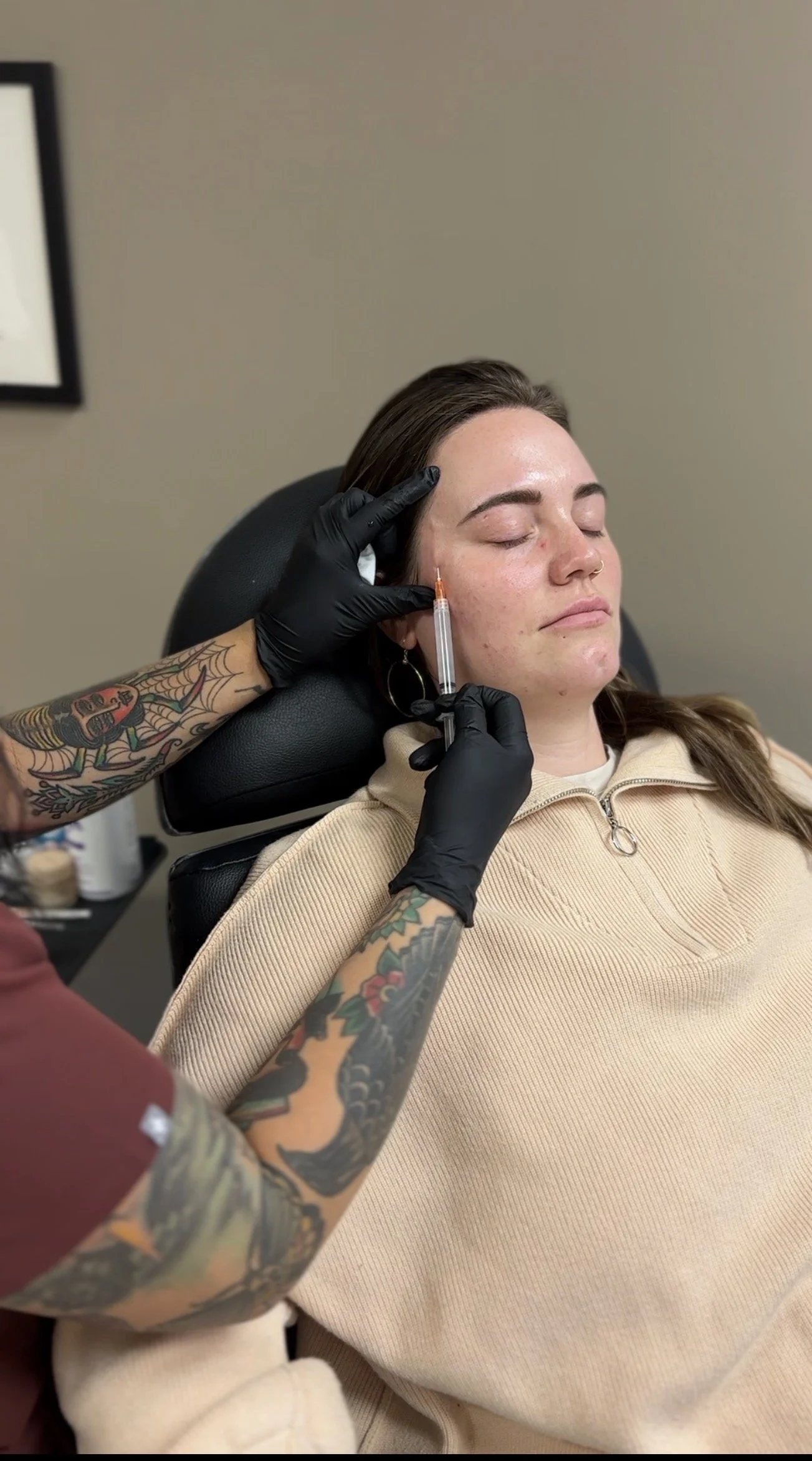 A woman with closed eyes receiving a cosmetic injection in her cheek from a tattooed medical professional wearing black gloves in a clinic.