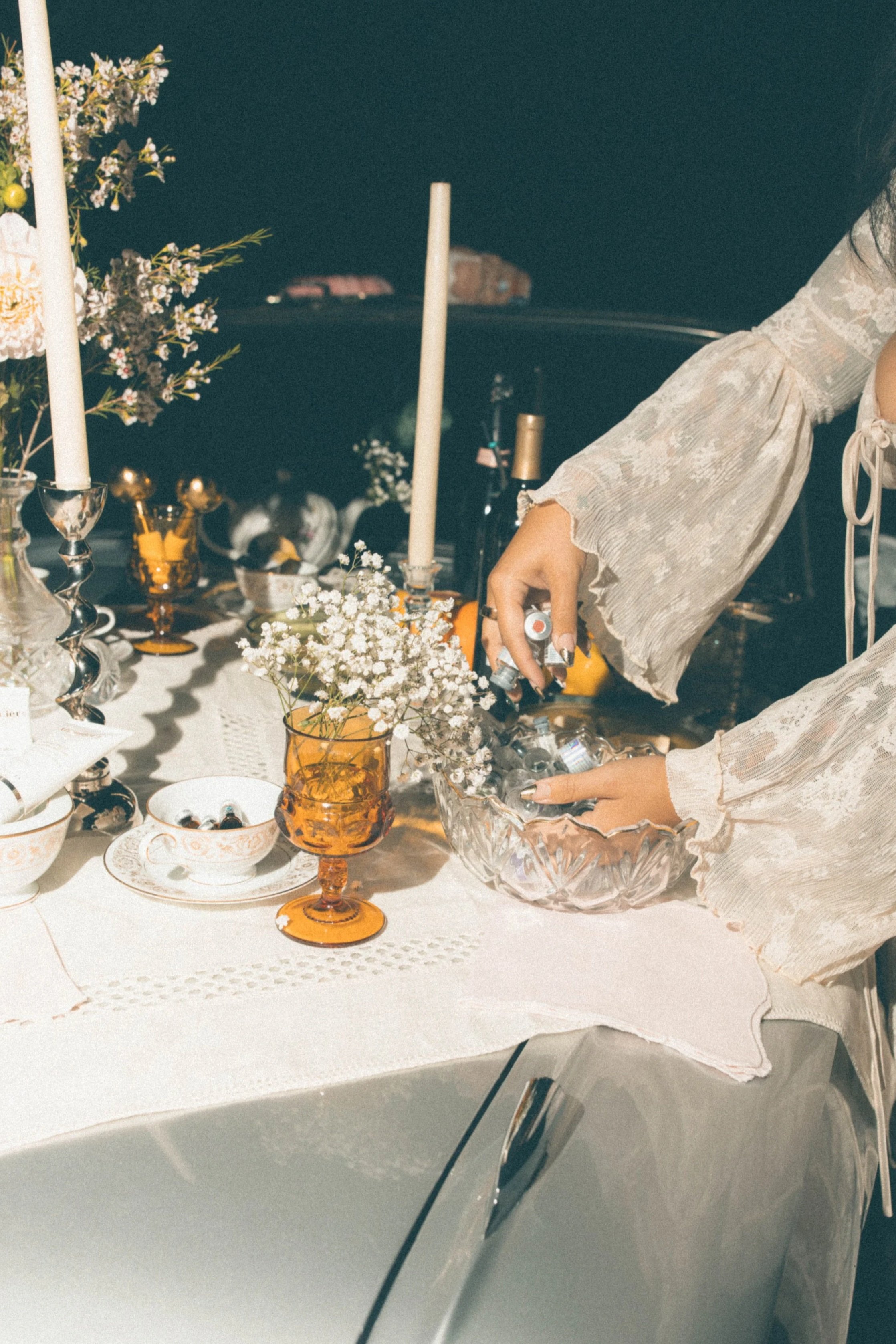 A woman in a lace long-sleeve dress reaches into a crystal bowl at a decorated table with candles, flowers, dishes, and glassware for a gathering or celebration.