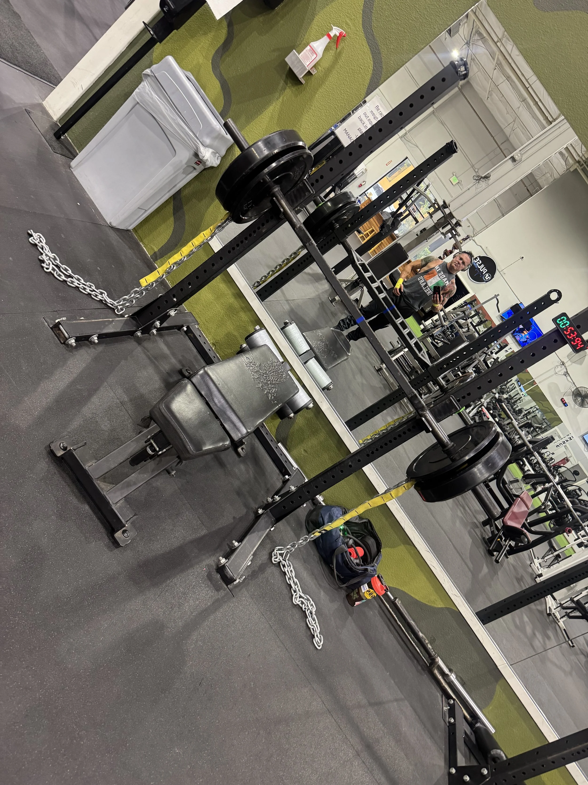 Three men posing in a martial arts gym, two are shirtless and wearing shorts, while the third is in a black T-shirt and pants. A cage and equipment are visible in the background.