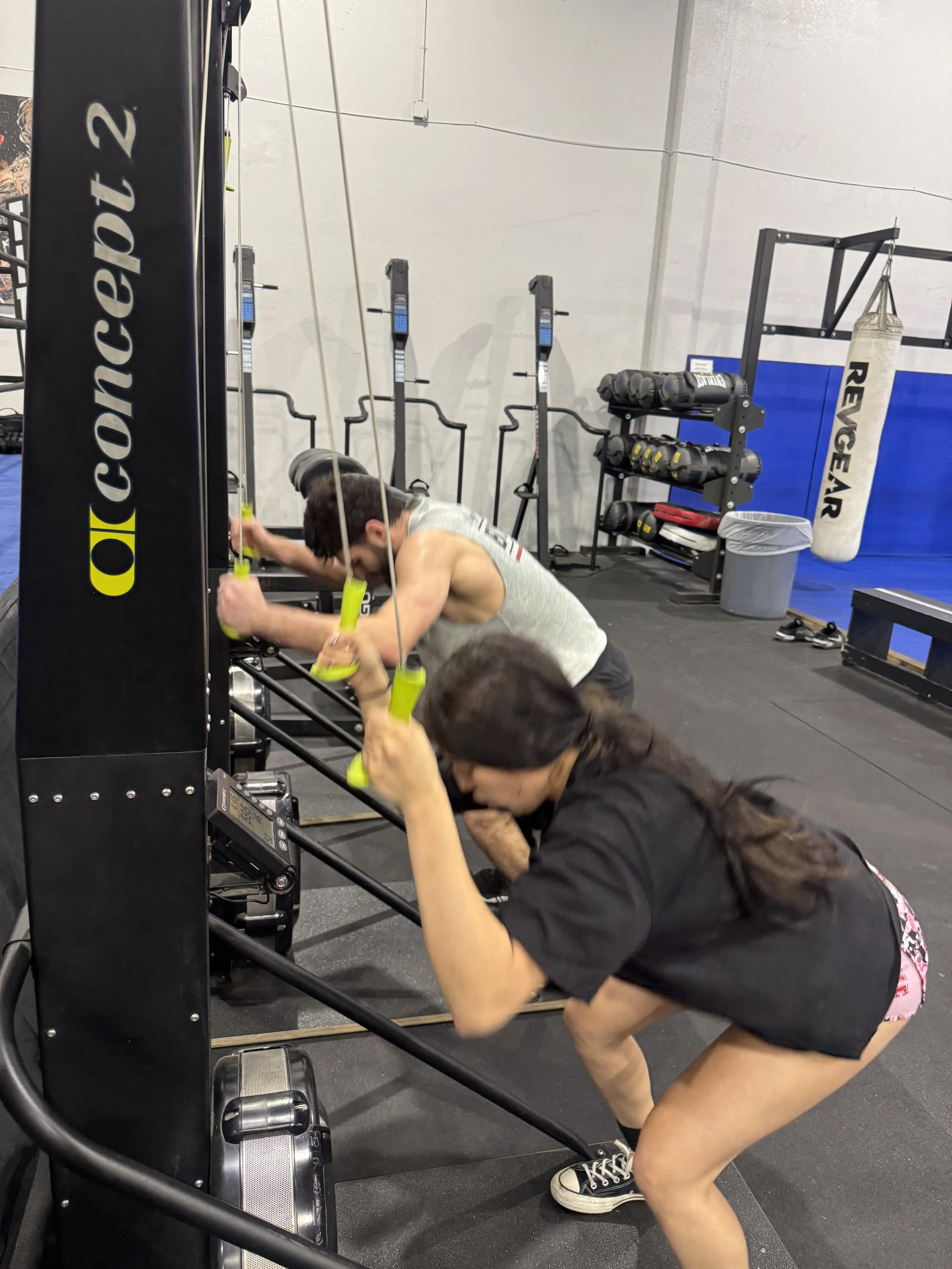 Two people exercising in a gym, one lifting dumbbells and the other standing with hands on hips. The gym has artificial turf and exercise equipment, with tall buildings visible outside.