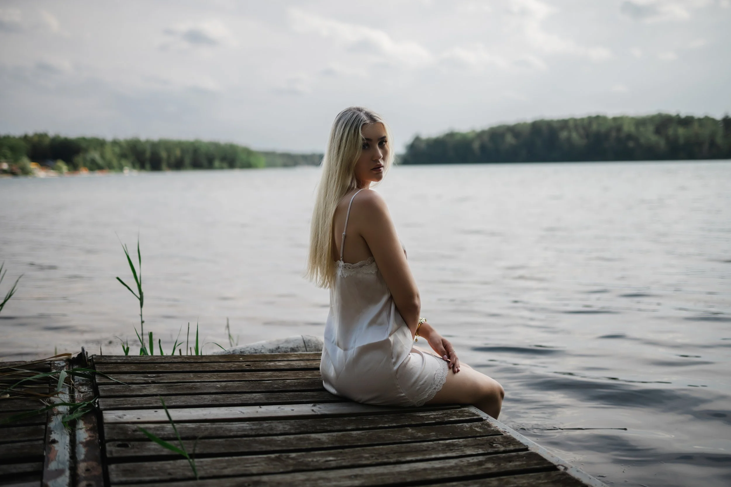  Sarina sitzt im hellen Satin-Kleid auf einem verwitterten Holzsteg am Steinberger See. 