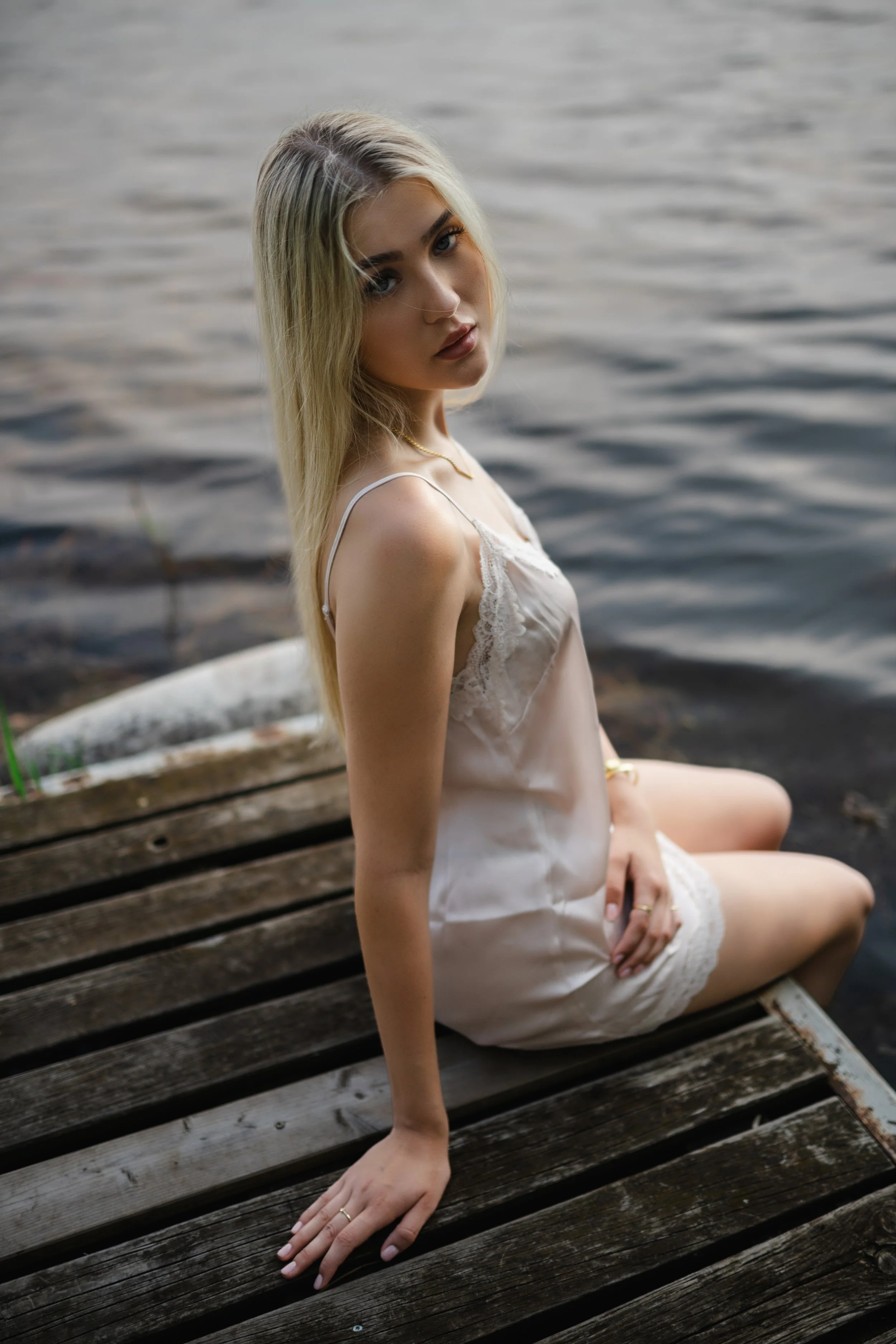  Sarina sitzt im hellen Satin-Kleid auf einem verwitterten Holzsteg am Steinberger See. 