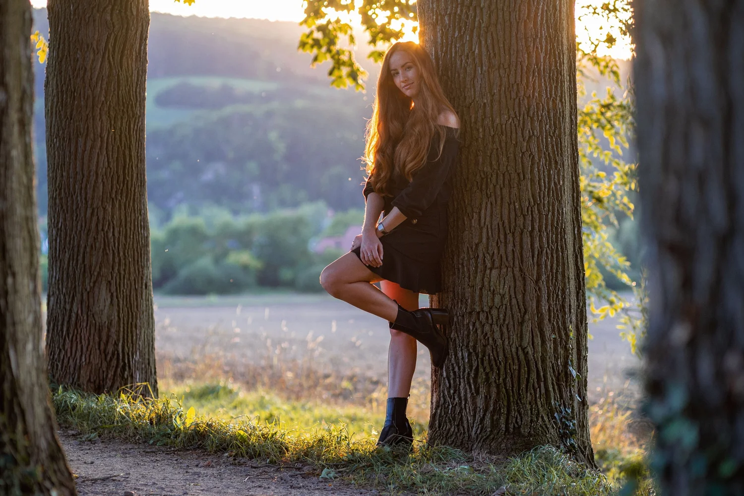 Gegenlichtportrait einer dunkel gekleideten Frau zwischen Baumstämmen vor einer hellen Landschaft