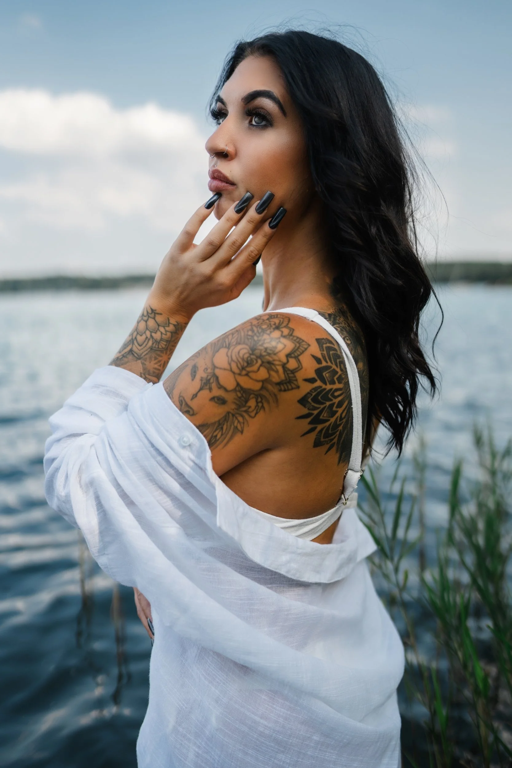 Portrait von Caro am See mit weißem Hemd, dunklem Haar und sichtbaren Tattoos.