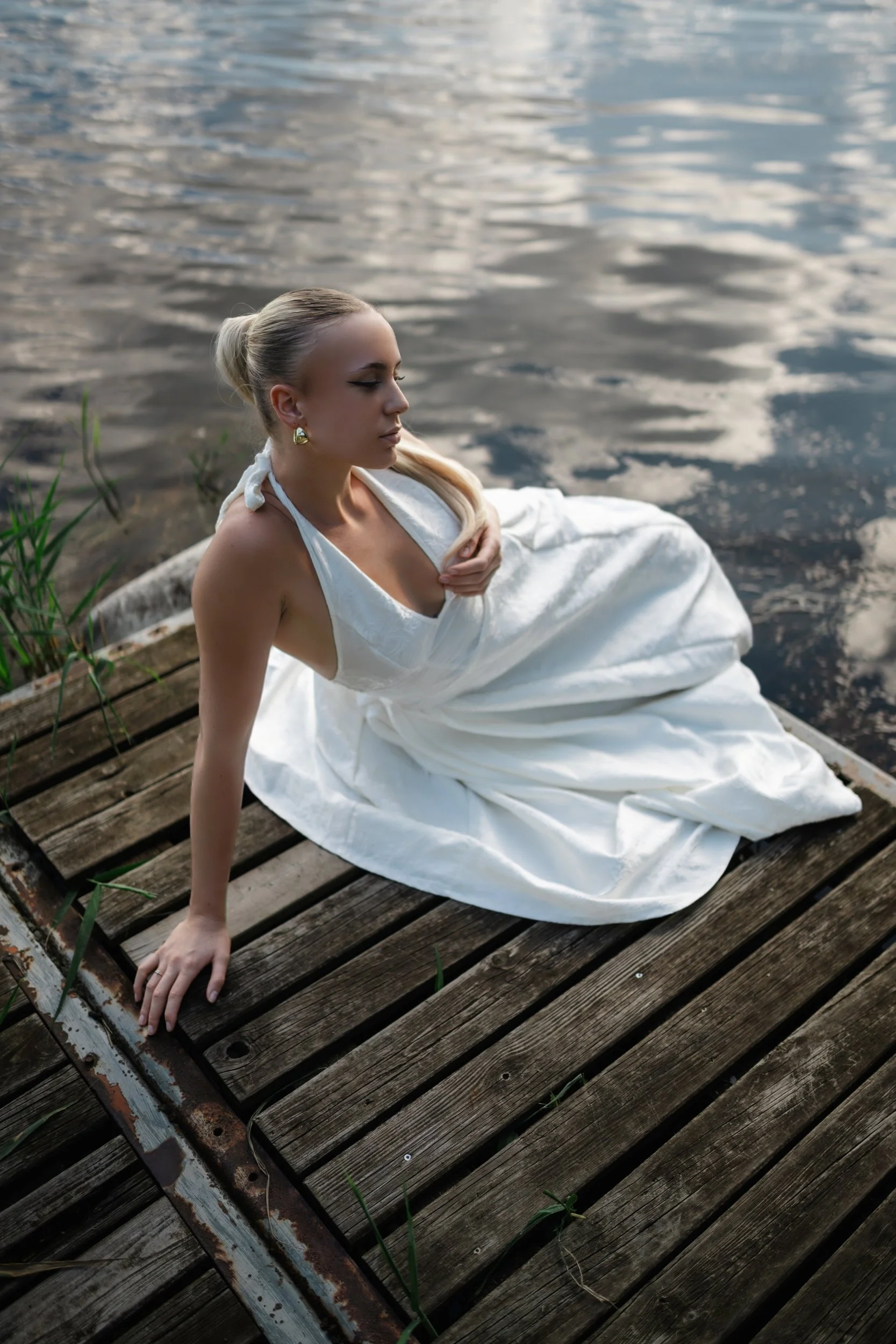 Sarah sitzt in einem weißen Kleid auf einem Holzsteg vor spiegelndem Wasser.