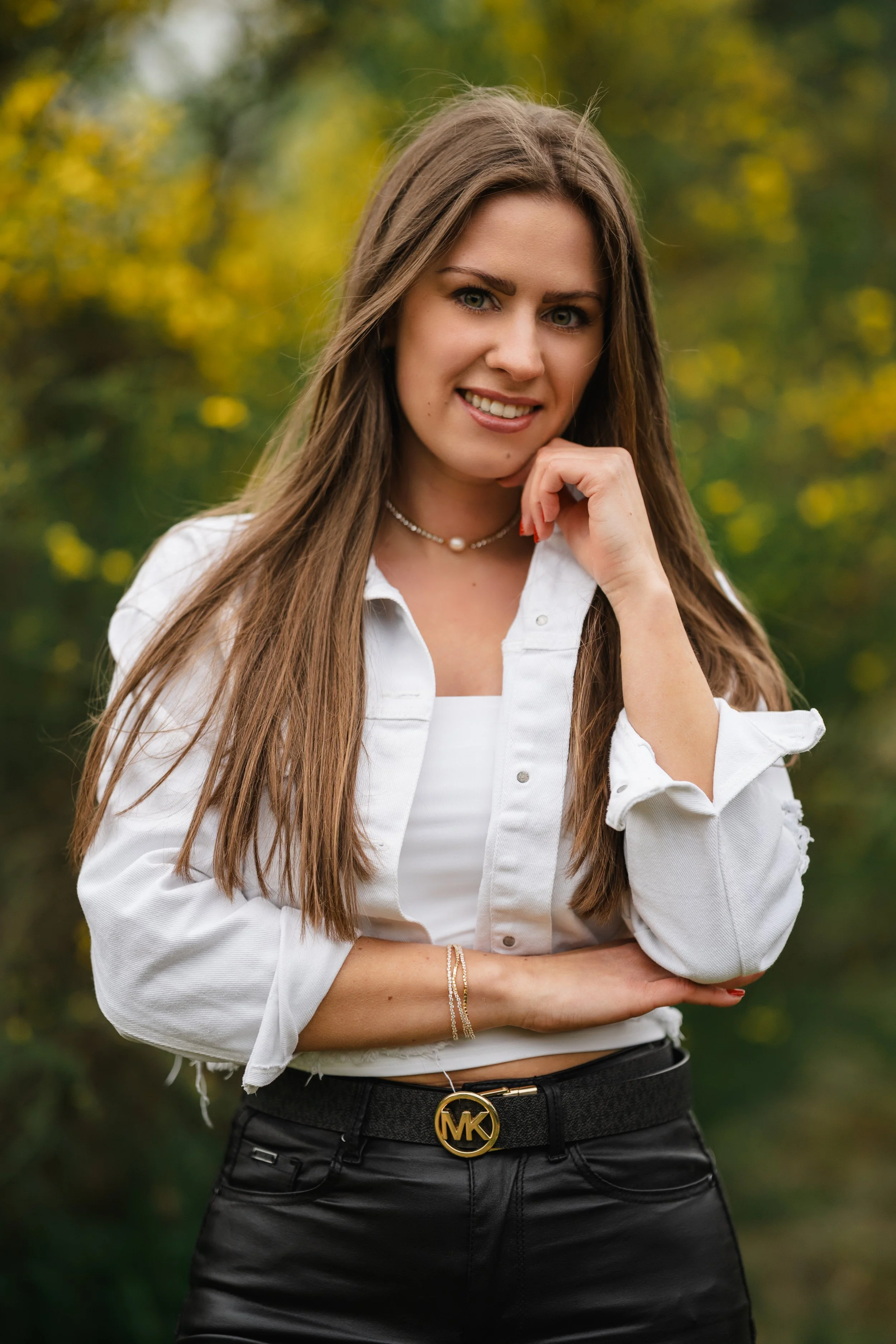 Sophia in weißer Bluse und schwarzer Hose vor unscharfem Grün mit gelben Blüten, beleuchtet von weichem Tageslicht.