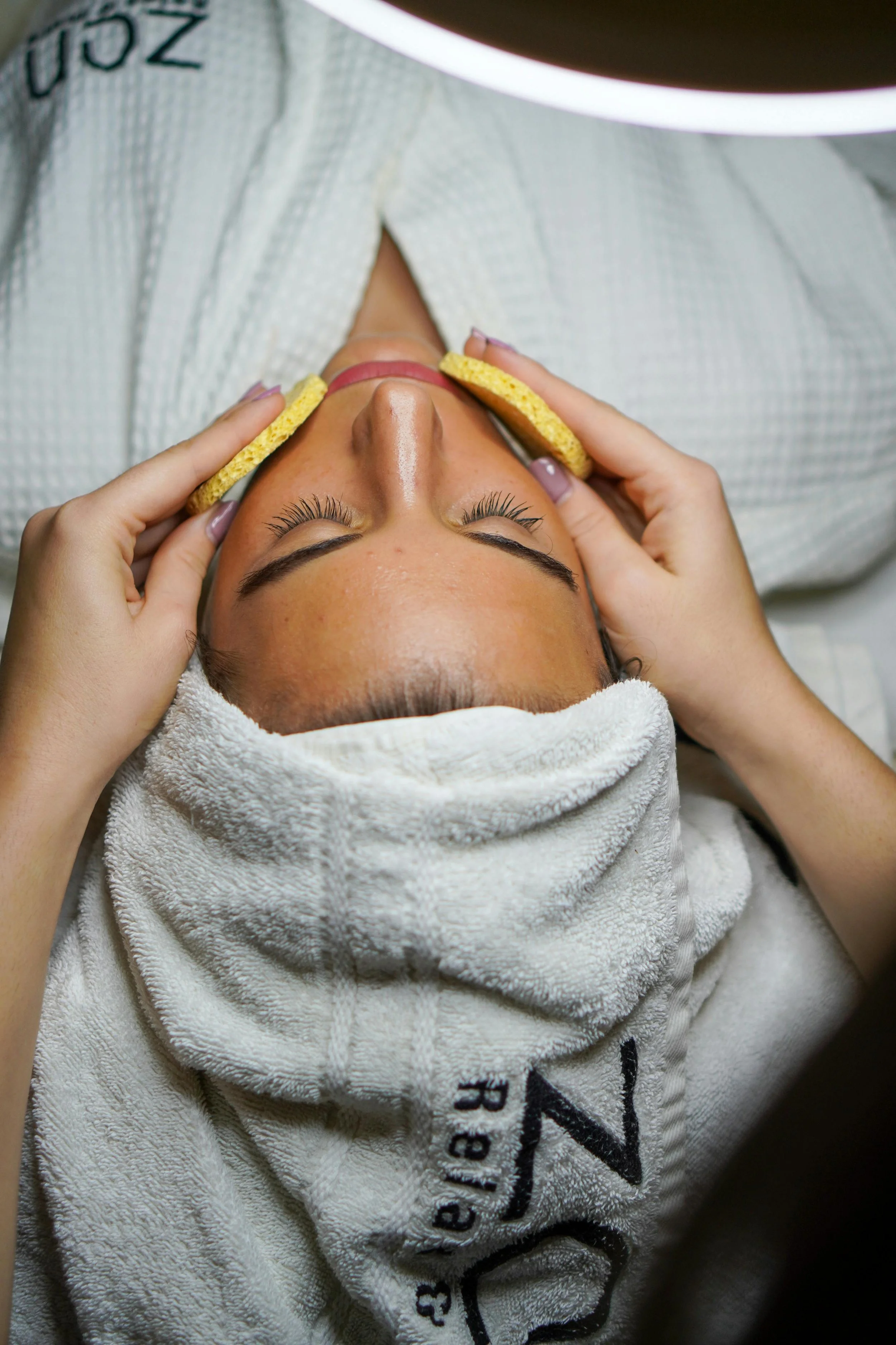 A woman receiving a facial massage with sponge on her face in a spa. She is lying down with a towel wrapped around her head and a white robe, in a relaxing setting.