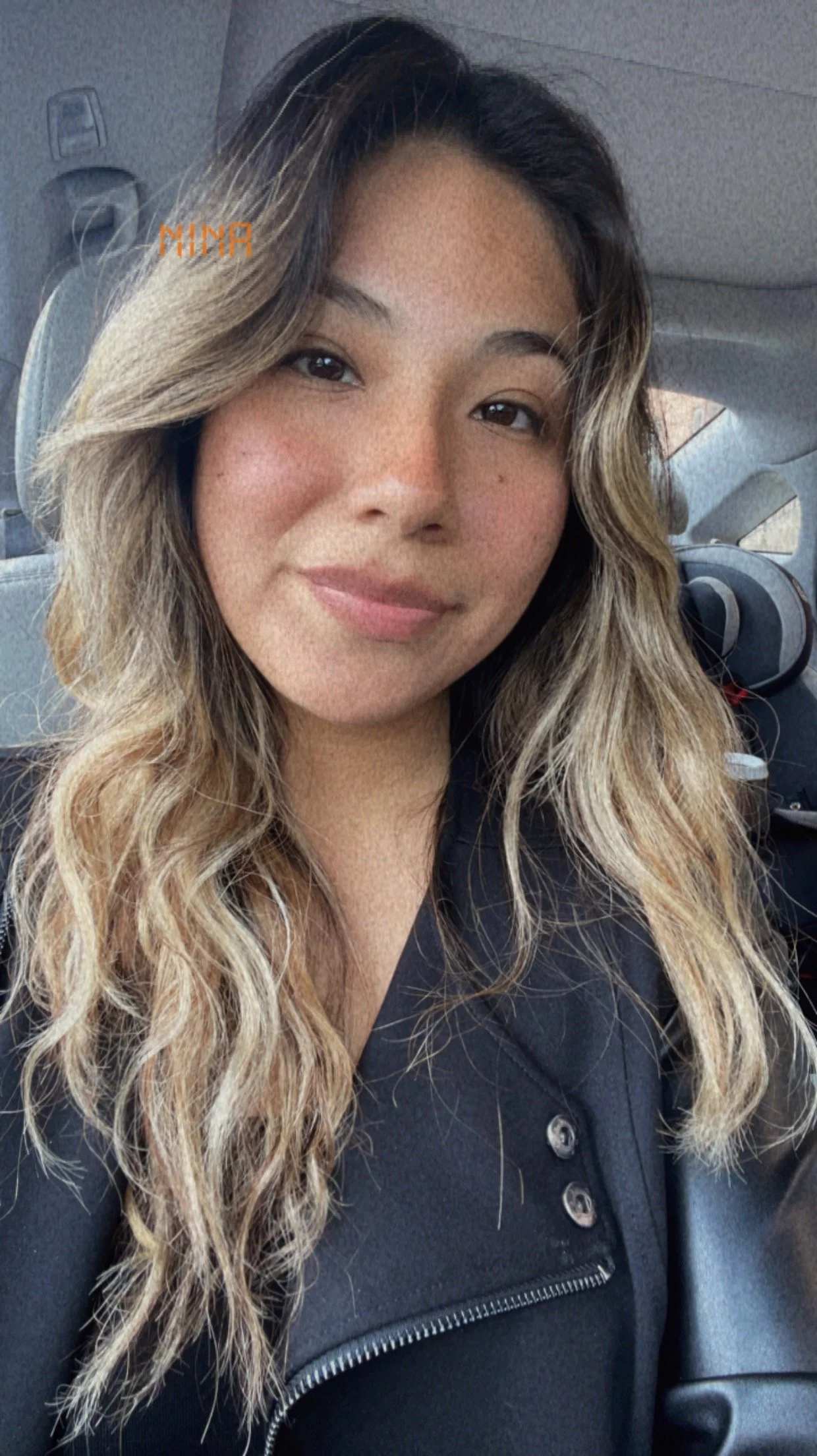 A woman with wavy light brown hair and freckles sitting inside a car, smiling at the camera.
