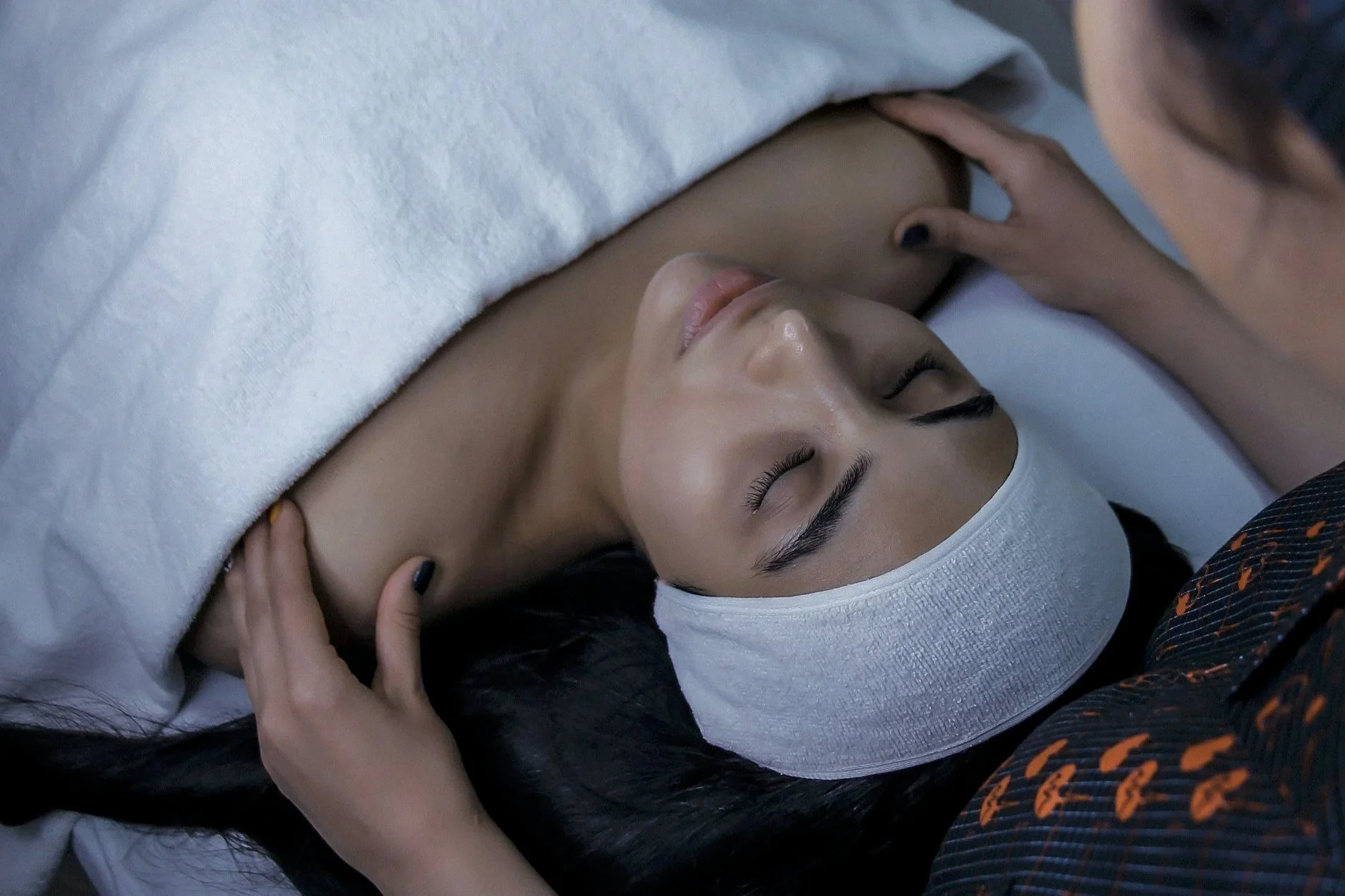 A woman receiving a massage while lying on a massage table with her eyes closed, wearing a white headband and covered with a white blanket.