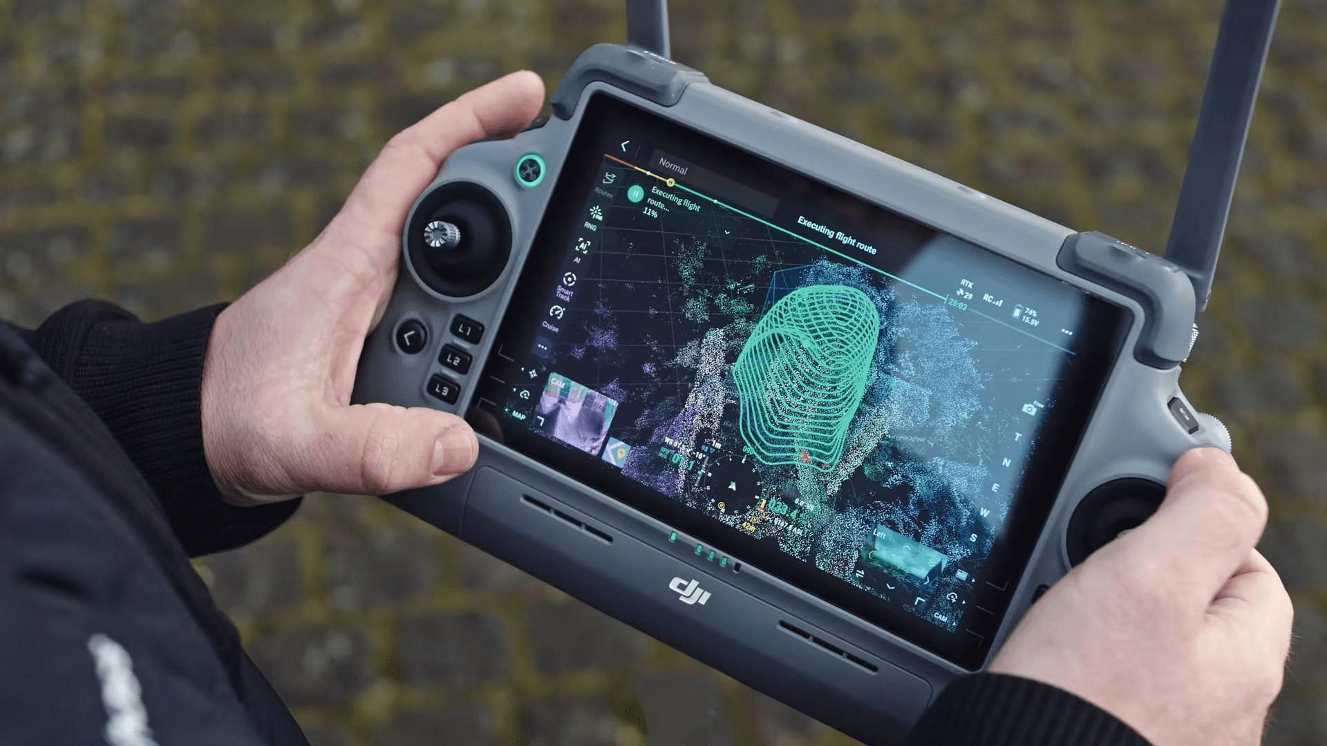 Person holding a DJI drone controller with a display screen showing a 3D map and flight route visualization in an outdoor setting with a cobblestone surface.