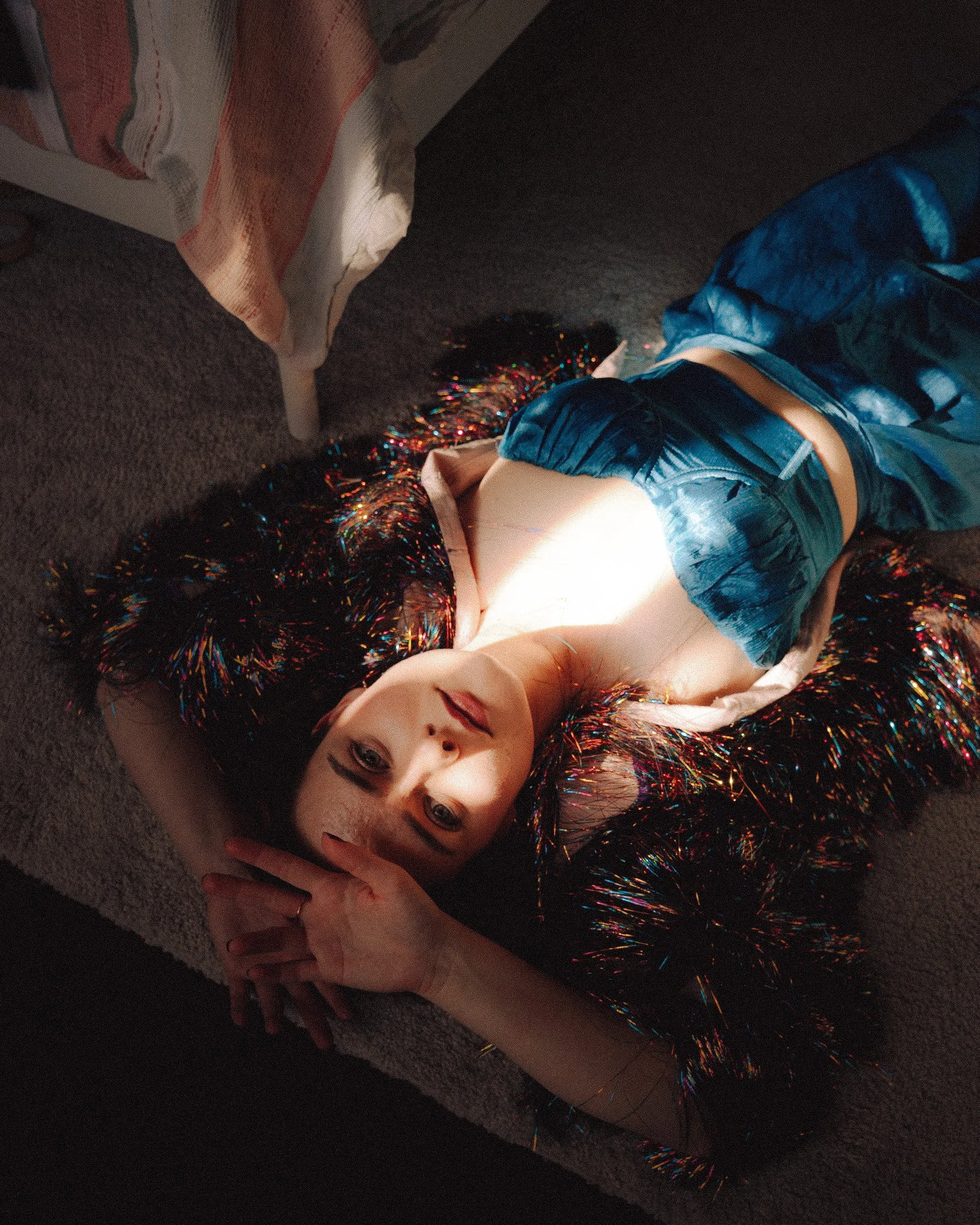 A young woman with dark hair and makeup, lying on the carpeted floor, looking at the camera. She's wearing a shiny, colorful jacket and blue satin pants, with a beige shirt underneath. A concentrated beam of light illuminates her face and chest, crea
