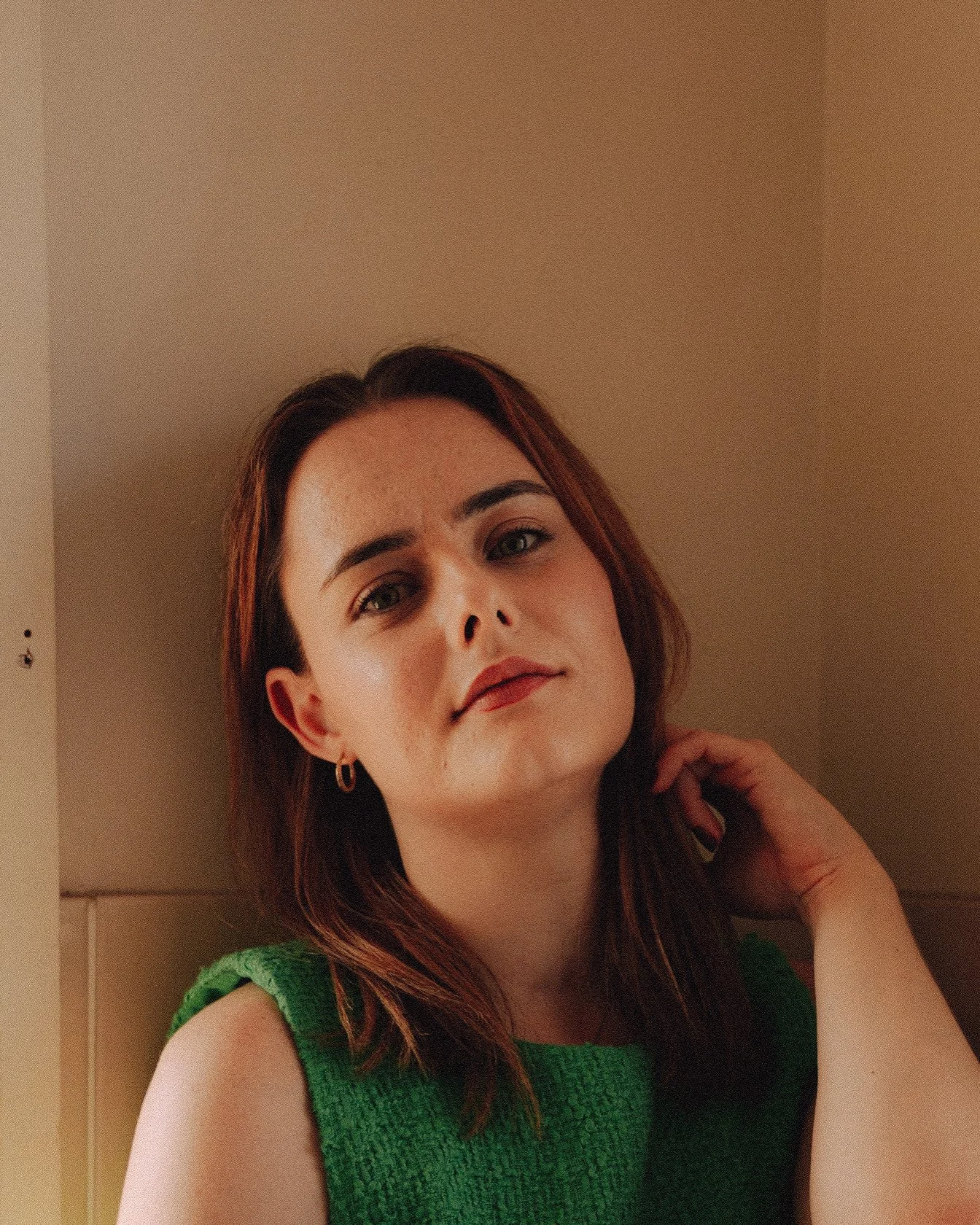 A woman with shoulder-length brown hair, wearing a green sleeveless top, poses against a beige wall with a slight tilt of her head, looking directly at the camera with a relaxed expression.