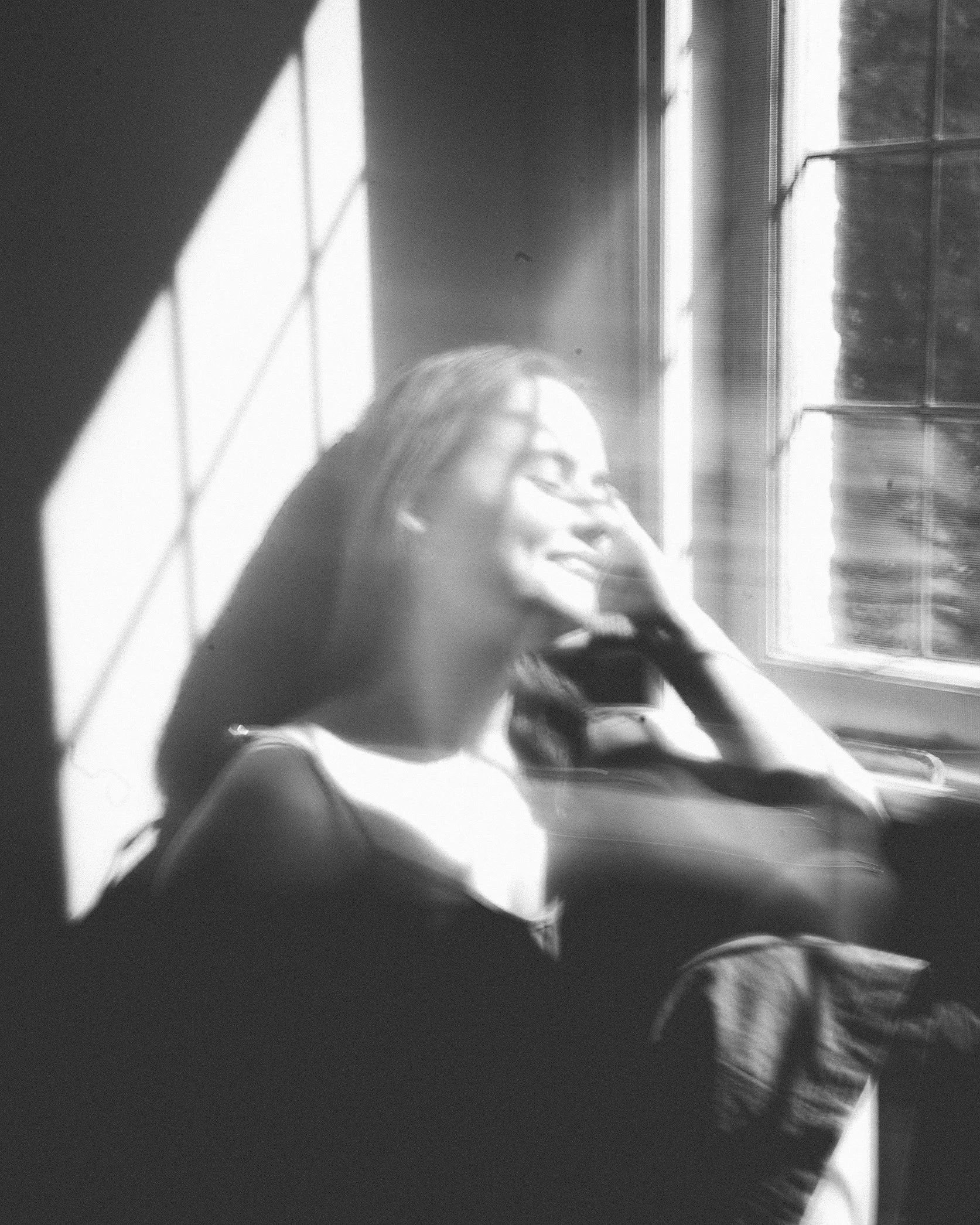A woman sitting by a window with sunlight streaming through, smiling and relaxing with her eyes closed, in a black and white photo.