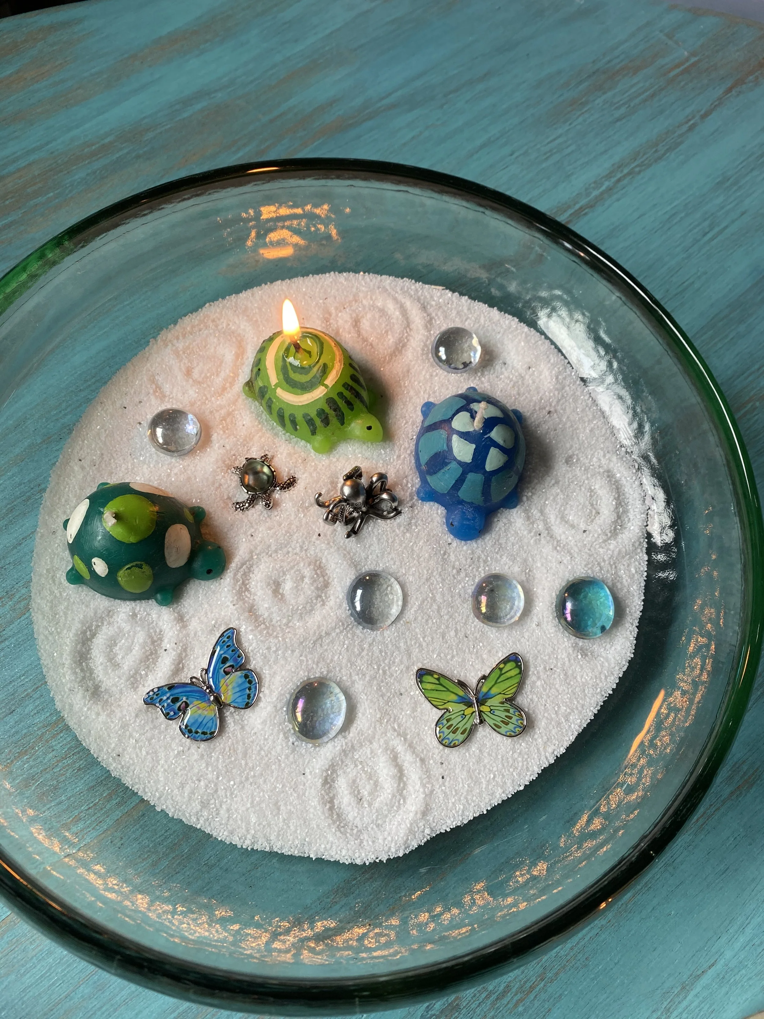 Decorative glass bowl filled with white sand, featuring colorful turtle-shaped candles, butterfly and ladybug charms, and small glass stones, with a lit green turtle candle.