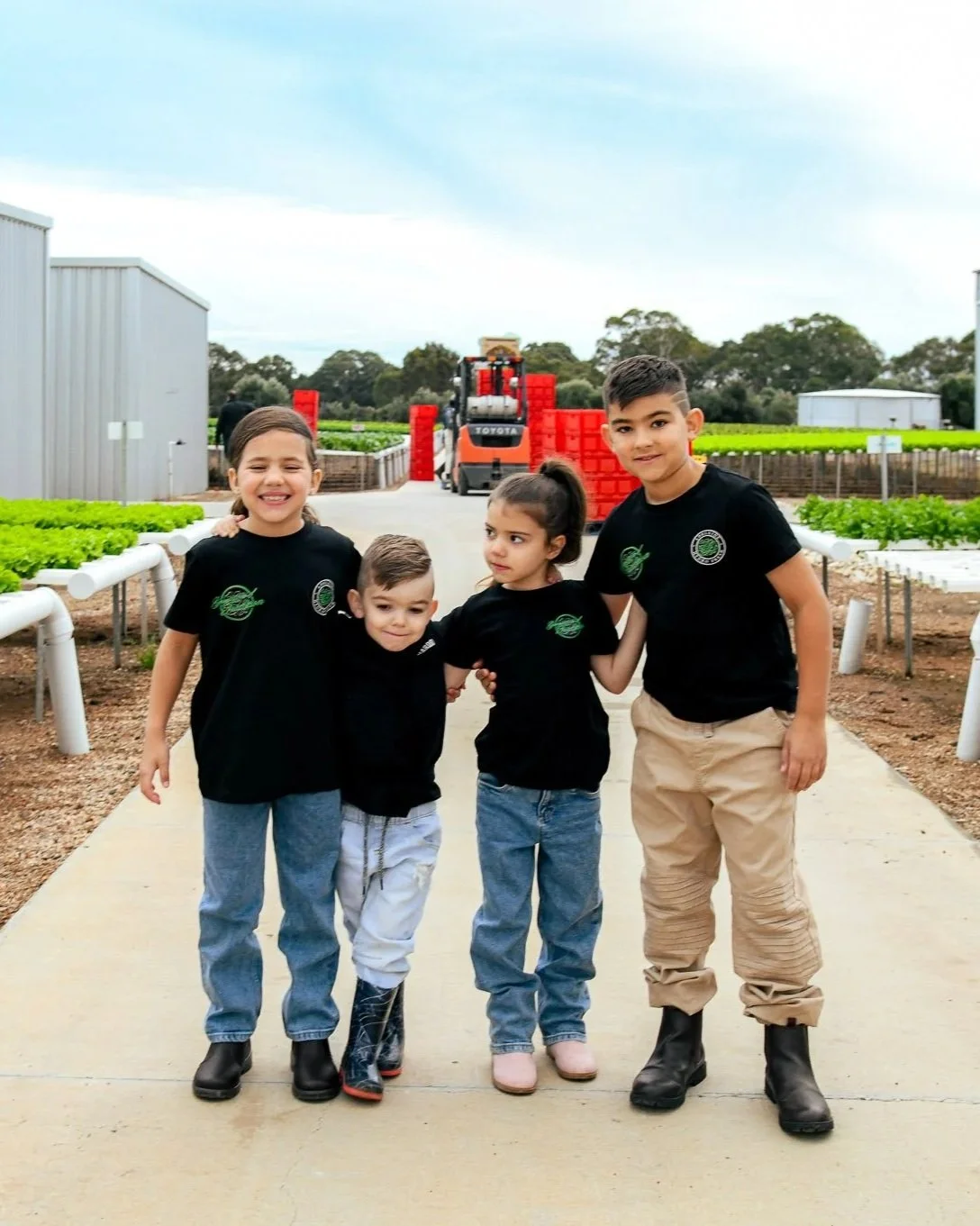 Four children standing on a farm or greenhouse with rows of plants and a forklift in the background, dressed in black shirts and casual pants.