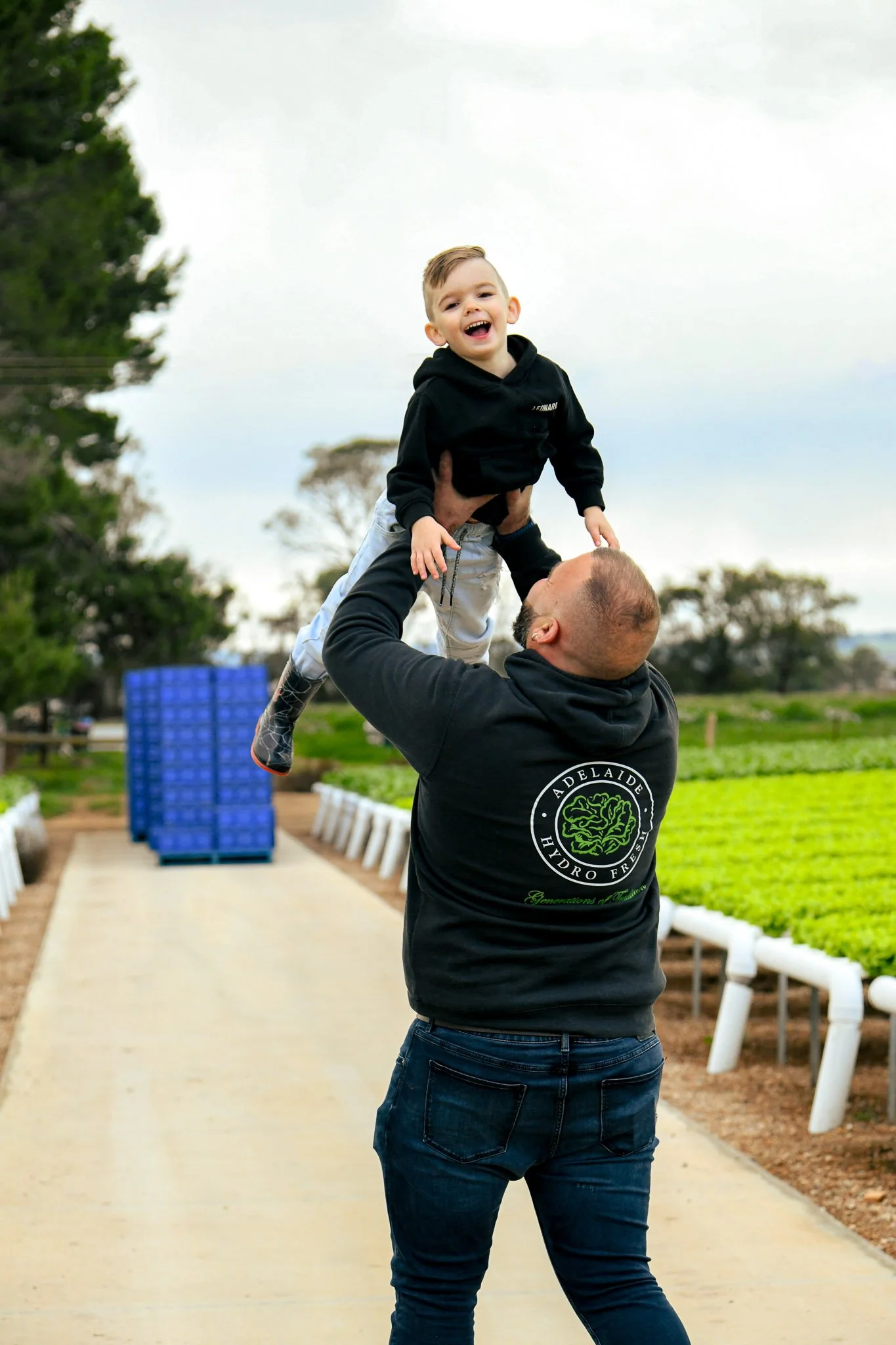 A man is lifting a smiling boy in the air outdoors, with greenery and farm or garden setting in the background.