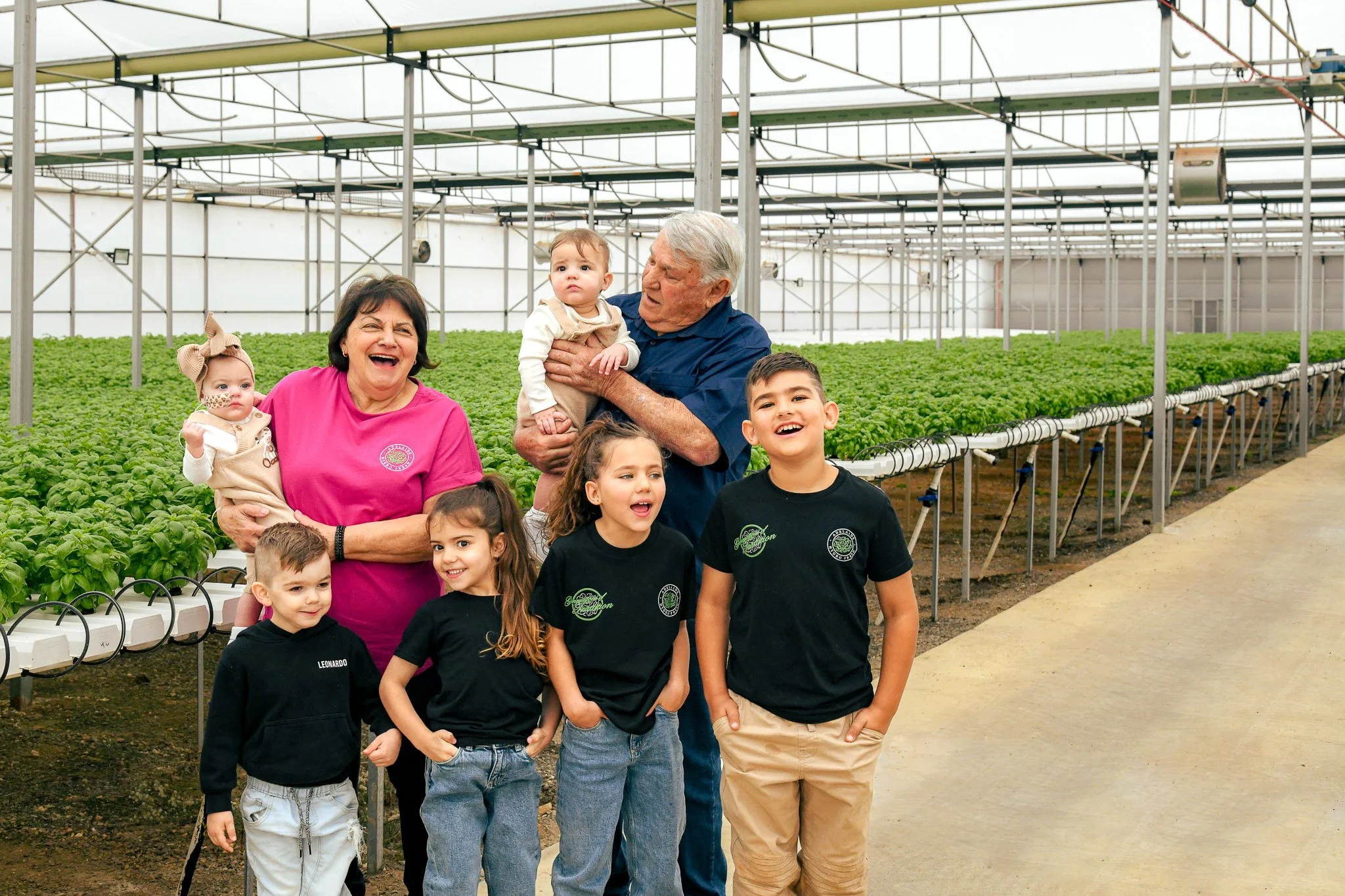 Adelaide Hydro Fresh: Grandparents and grandchildren standing in a greenhouse with green plants growing in rows. The group is smiling and appears happy.