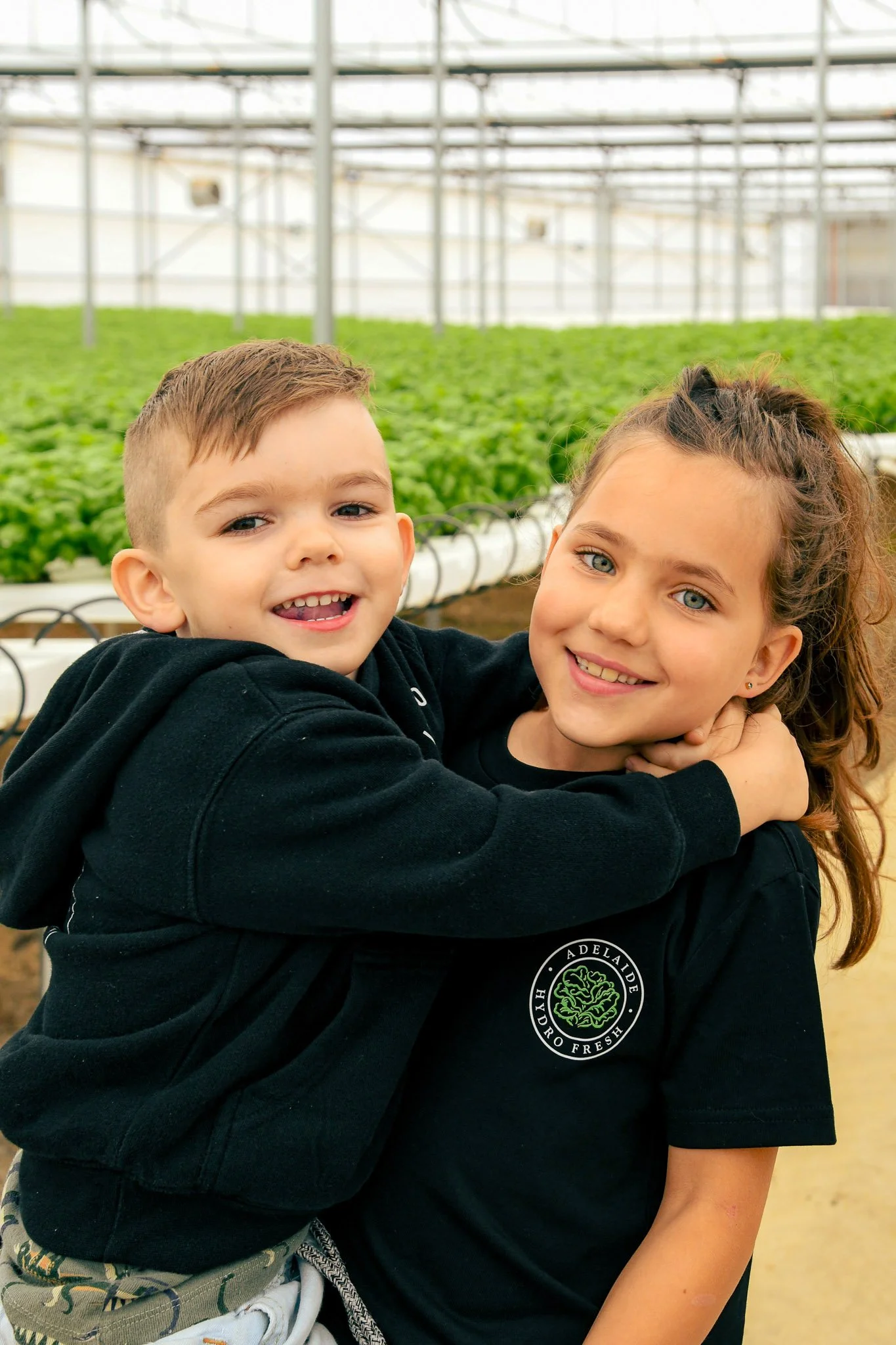 Two children, a boy and a girl, are hugging each other in a greenhouse with green plants in the background. They are both smiling and wearing black hoodies.