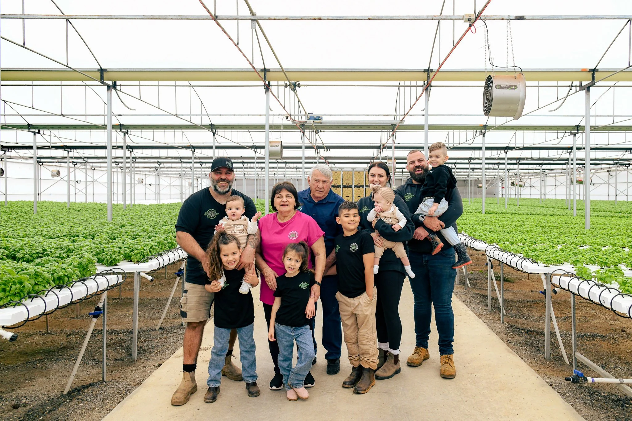 Adelaide Hydro Fresh family, standing inside a large greenhouse with rows of green plants on hydroponic systems.