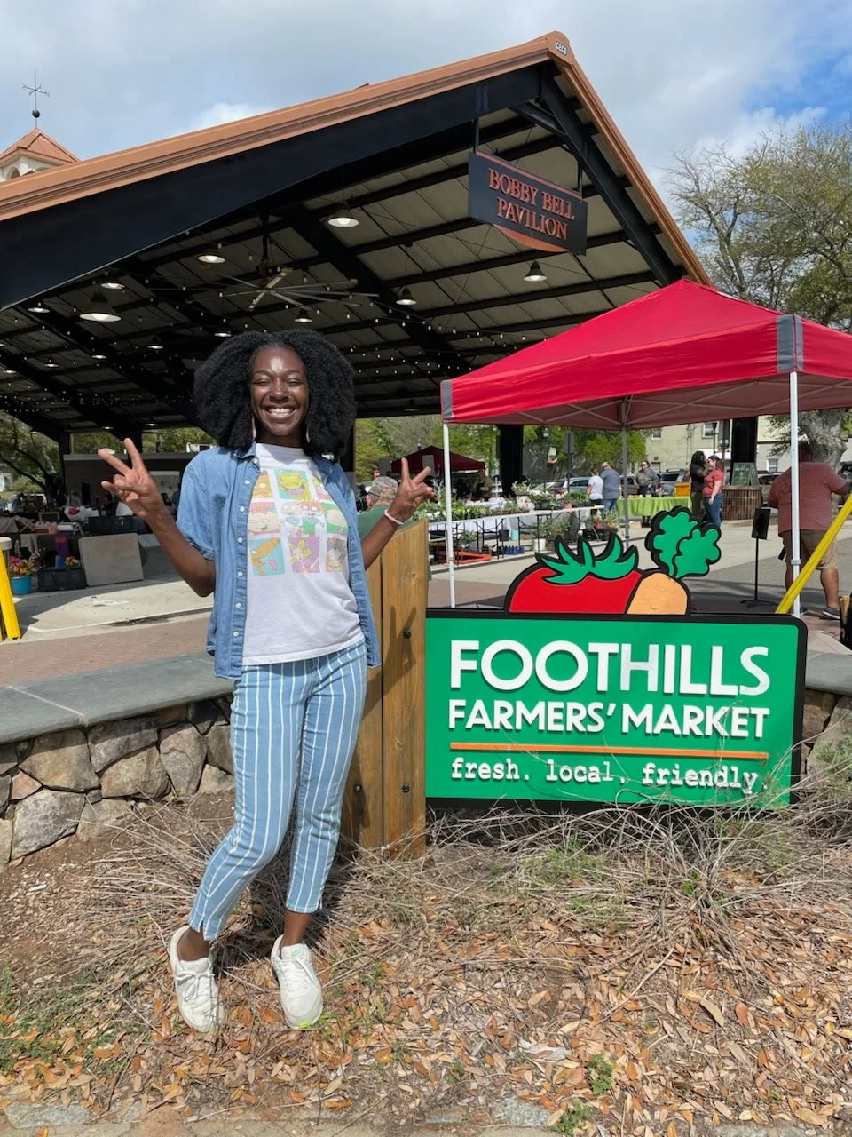 A young woman in casual dress standing and giving peace signs next to a foothills farmers' market sign