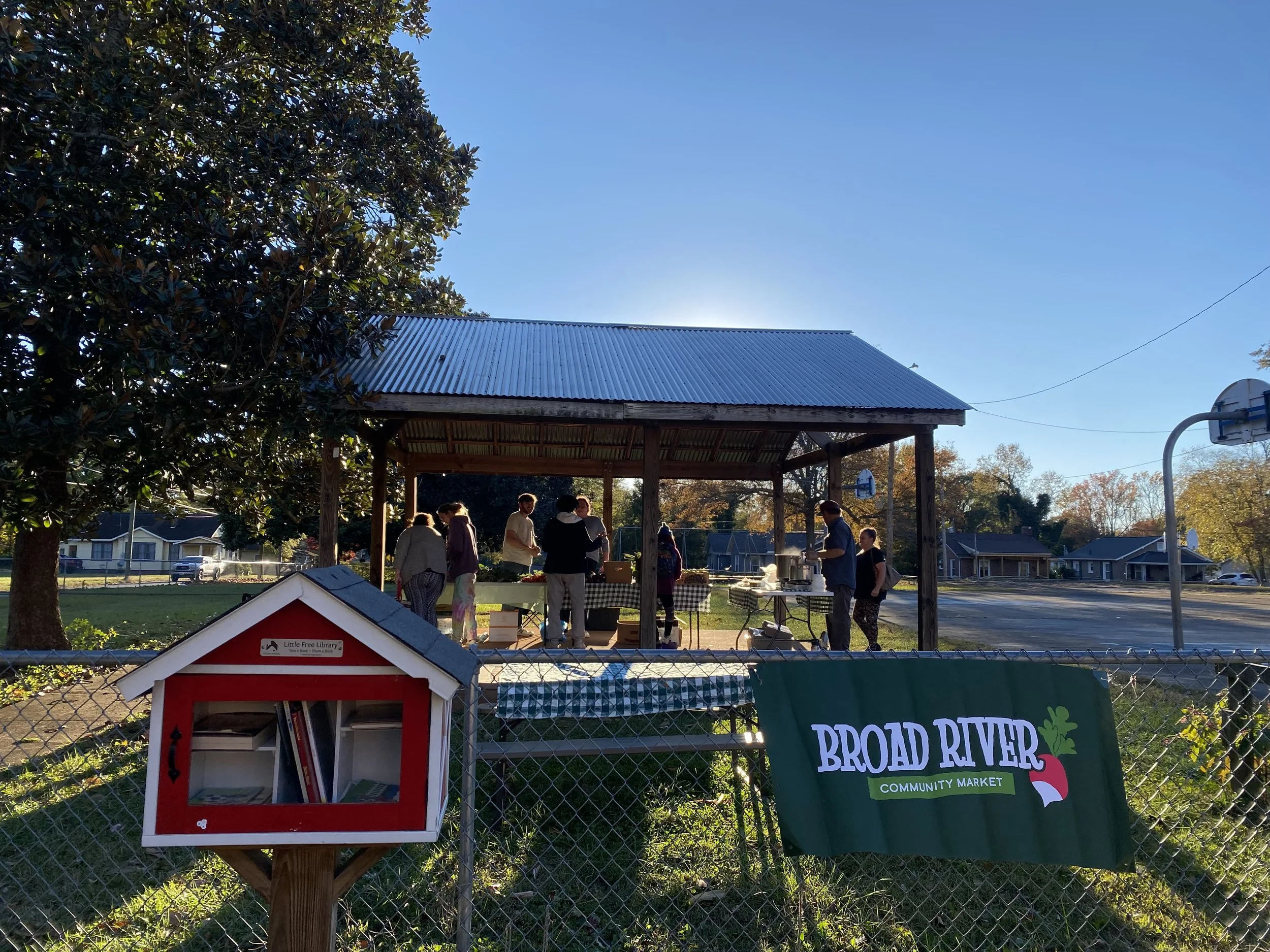 Community gathering at Broad River Community Market under a wooden pavilion with people talking and serving food. In the foreground, a red little free library box and a green Broad River Community Market banner are visible. There are residential houses and a basketball hoop in the background on a sunny day.