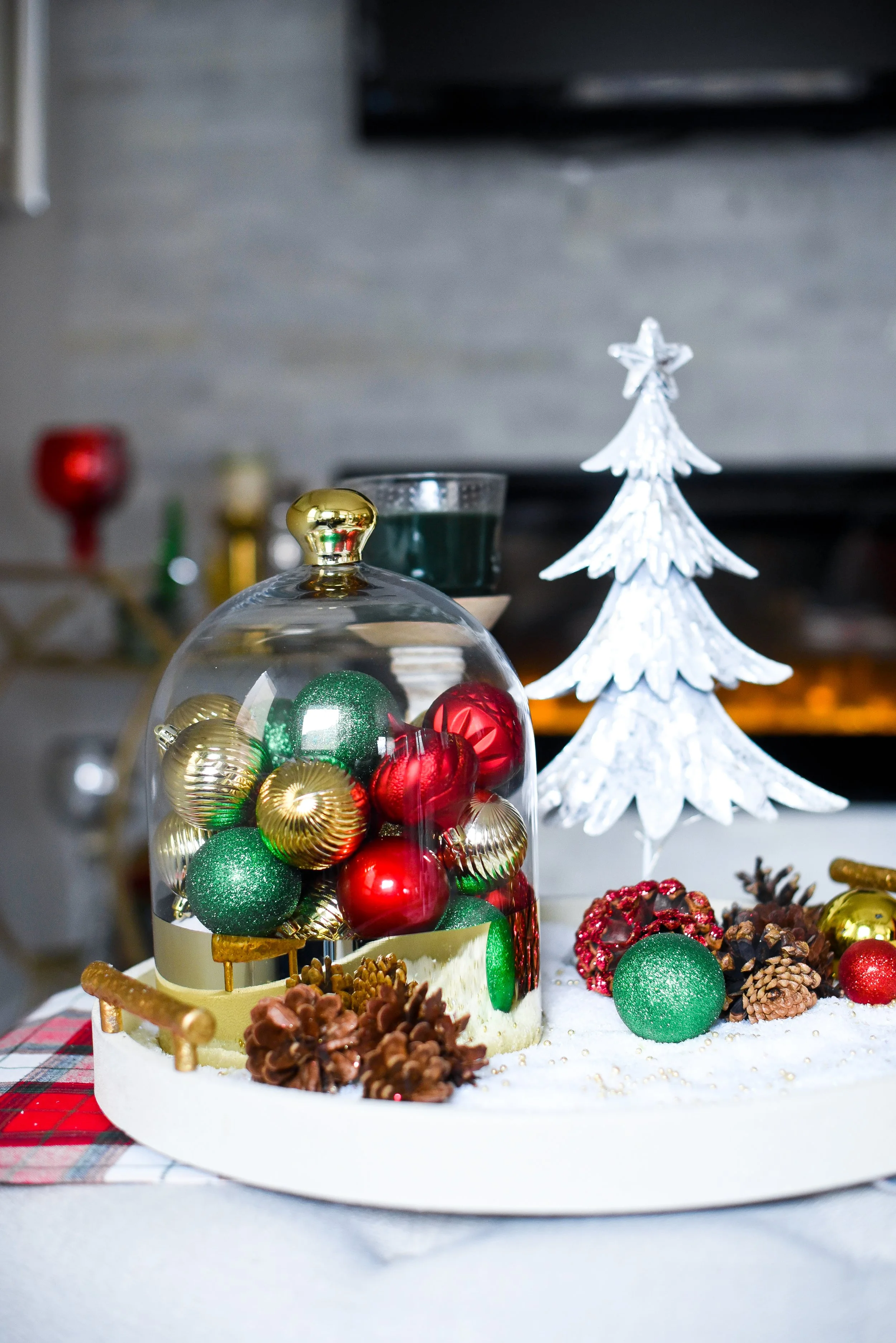 Christmas decorations on a table, including a glass dome filled with red, gold, and green ornaments, a white Christmas tree figurine, pinecones, and additional ornaments on a snowy surface.