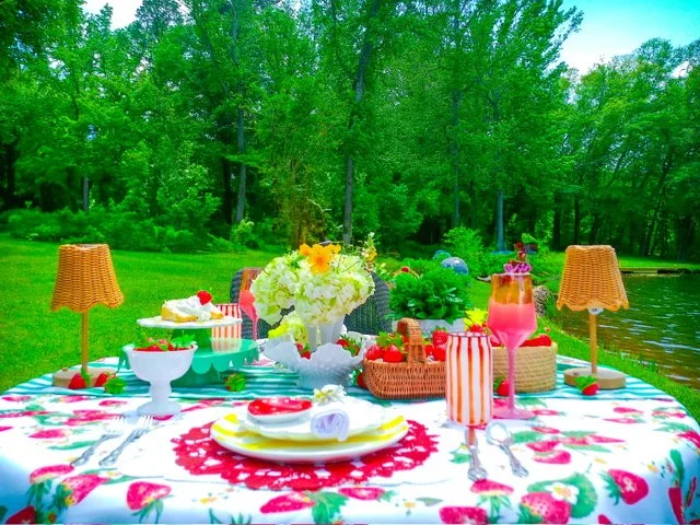 A colorful outdoor picnic table set on a floral tablecloth with plates, bowls, and drinks, surrounded by green trees and a body of water.