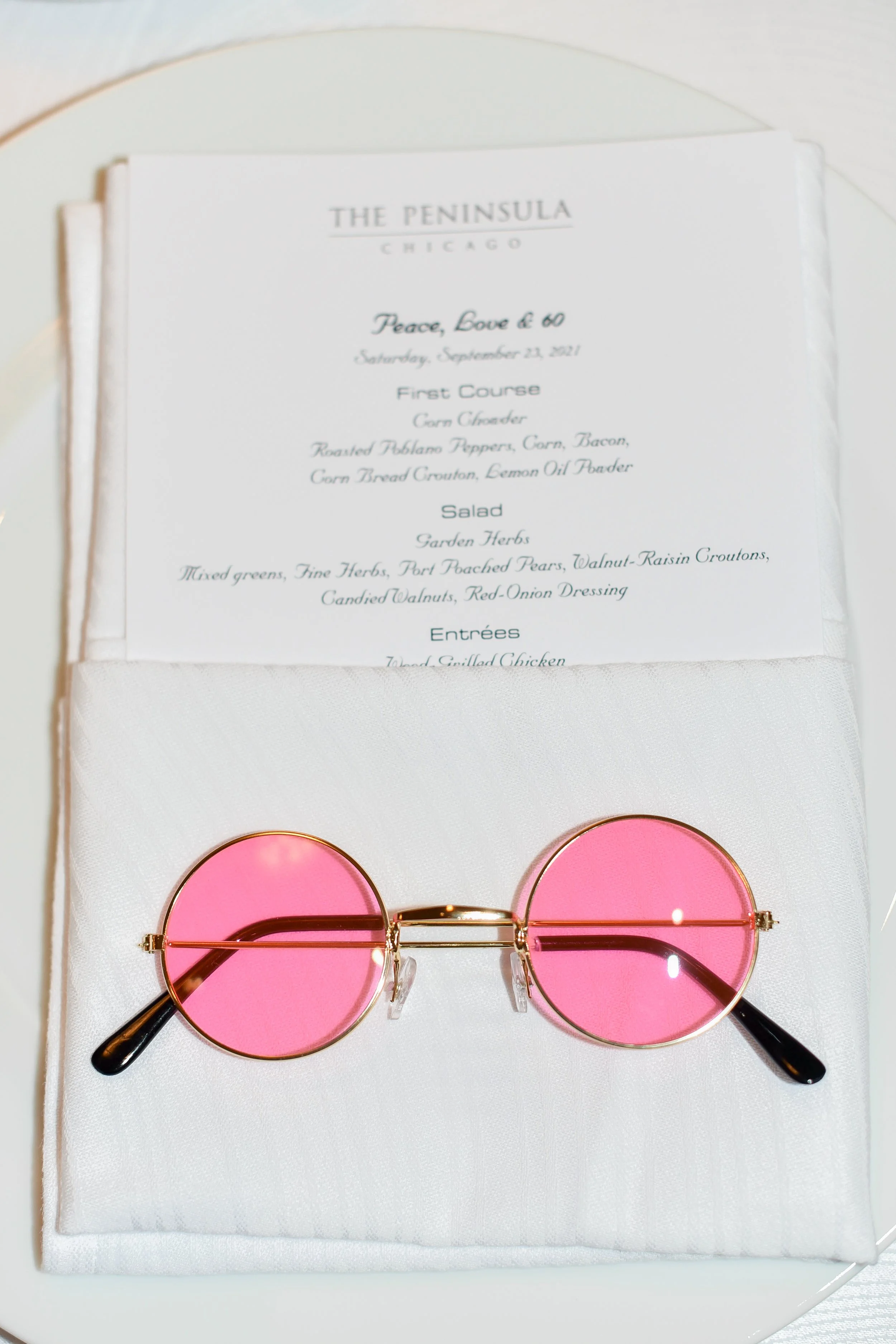 A pair of pink-tinted round sunglasses resting on a white cloth beneath a printed menu from The Peninsula Chicago restaurant.