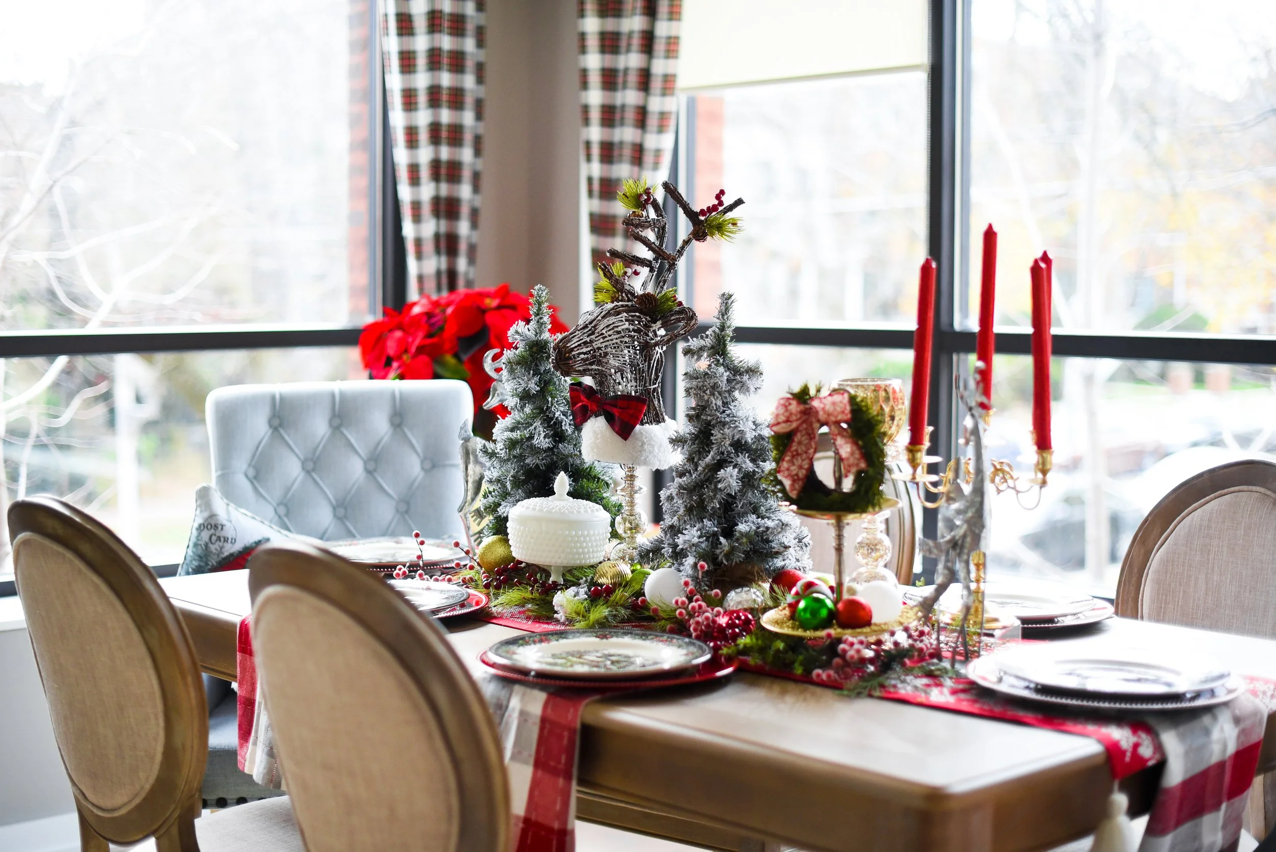 Decorated dining table with Christmas ornaments, a faux reindeer, small Christmas trees with snow, red candles, and festive greenery, set near large windows.