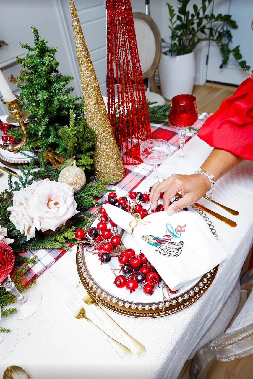 A festive holiday table decorated with small Christmas trees, gold and red cone-shaped decorations, greenery, and a plaid table runner. There are gold and red glasses, a hand with a bracelet, and a plate with a napkin featuring a colorful illustratio
