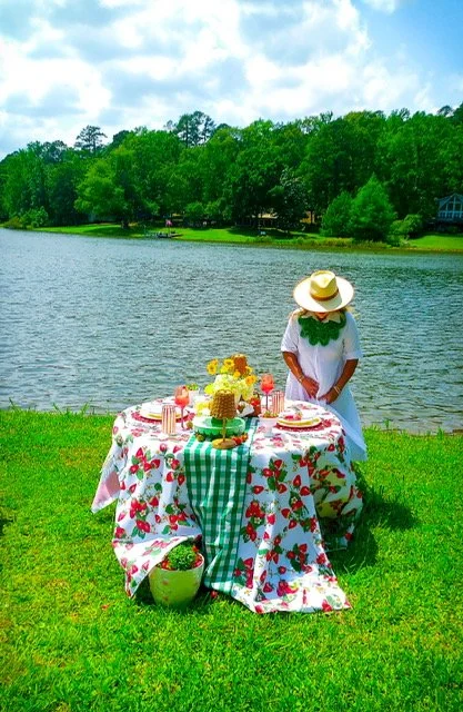 A woman dressed in white with a large straw hat standing near a table set for a picnic by a lake, with a green park and trees in the background.