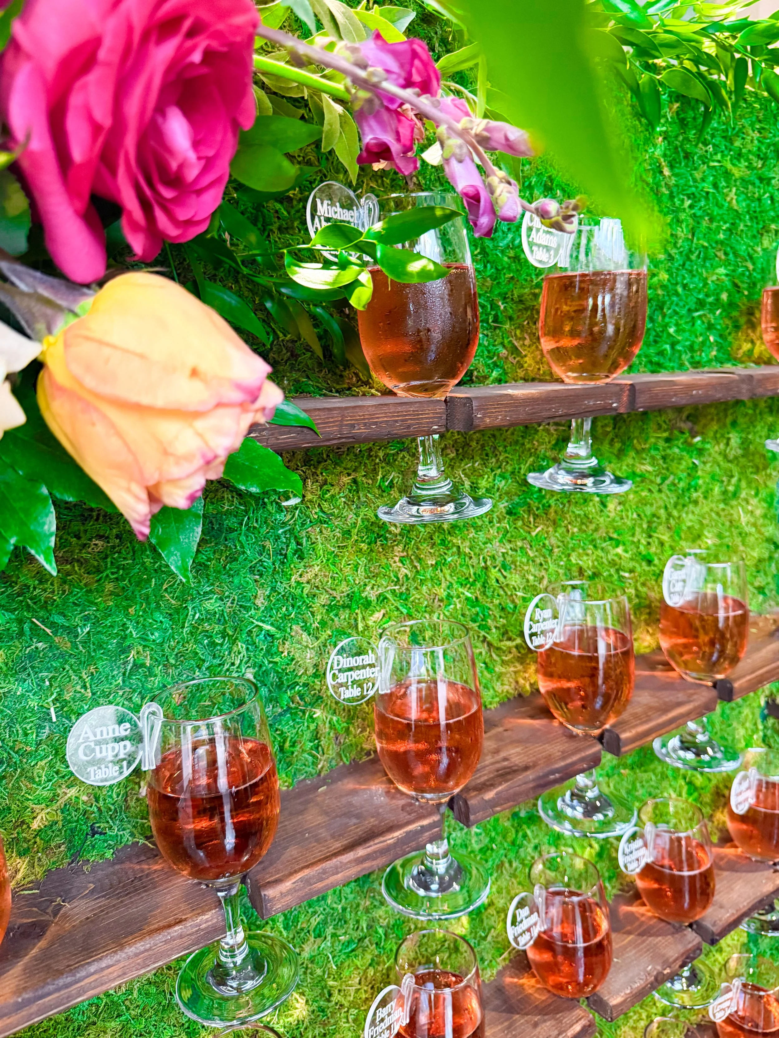 Glass jars filled with pinkish-orange liquid arranged on wooden shelves against a green, mossy background, decorated with colorful flowers and floral tags.
