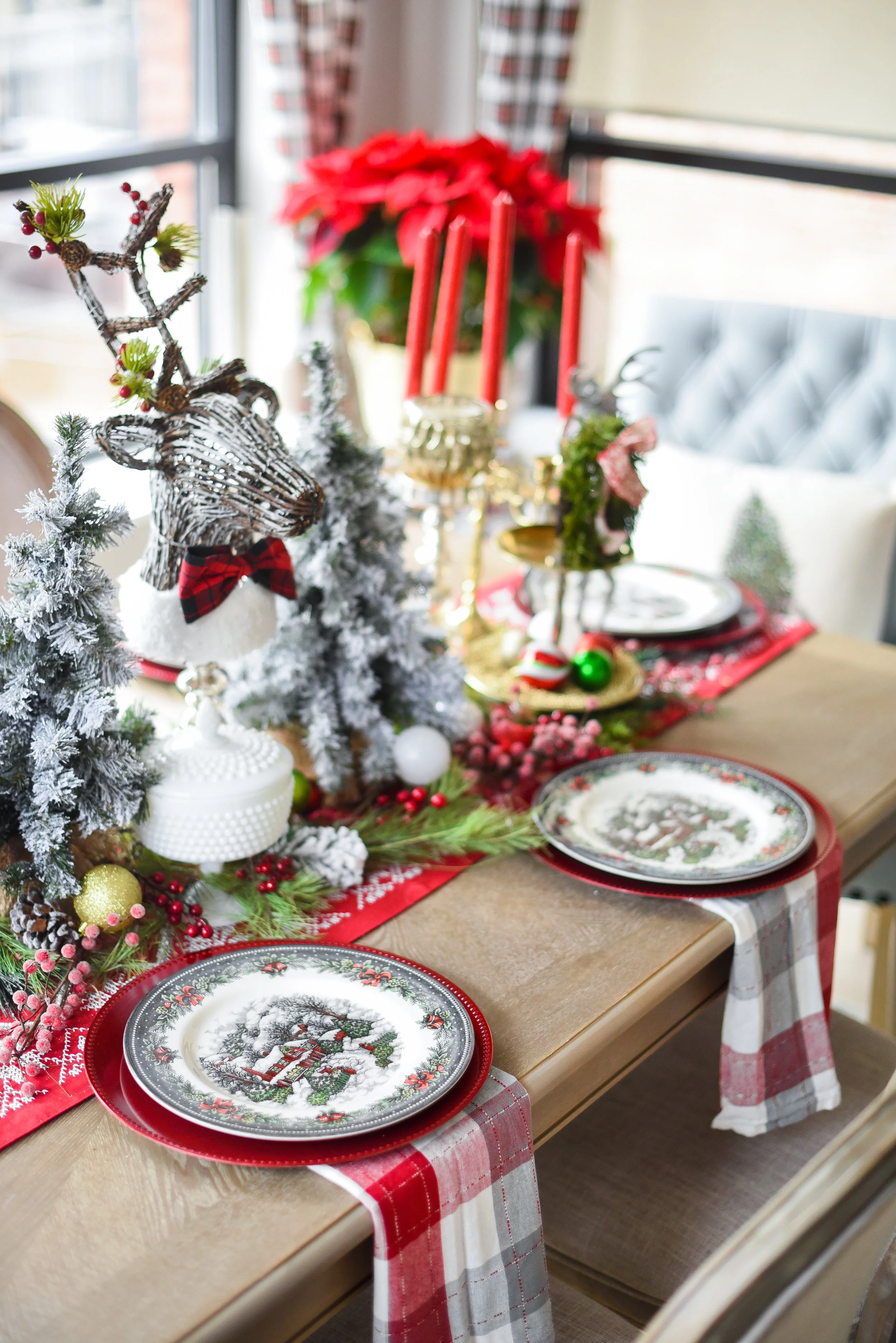 Decorated Christmas dining table with Christmas plates, holiday centerpiece featuring miniature snow-covered trees, reindeer figurine wearing a bow tie, and festive ornaments, with a poinsettia and red candles in the background.