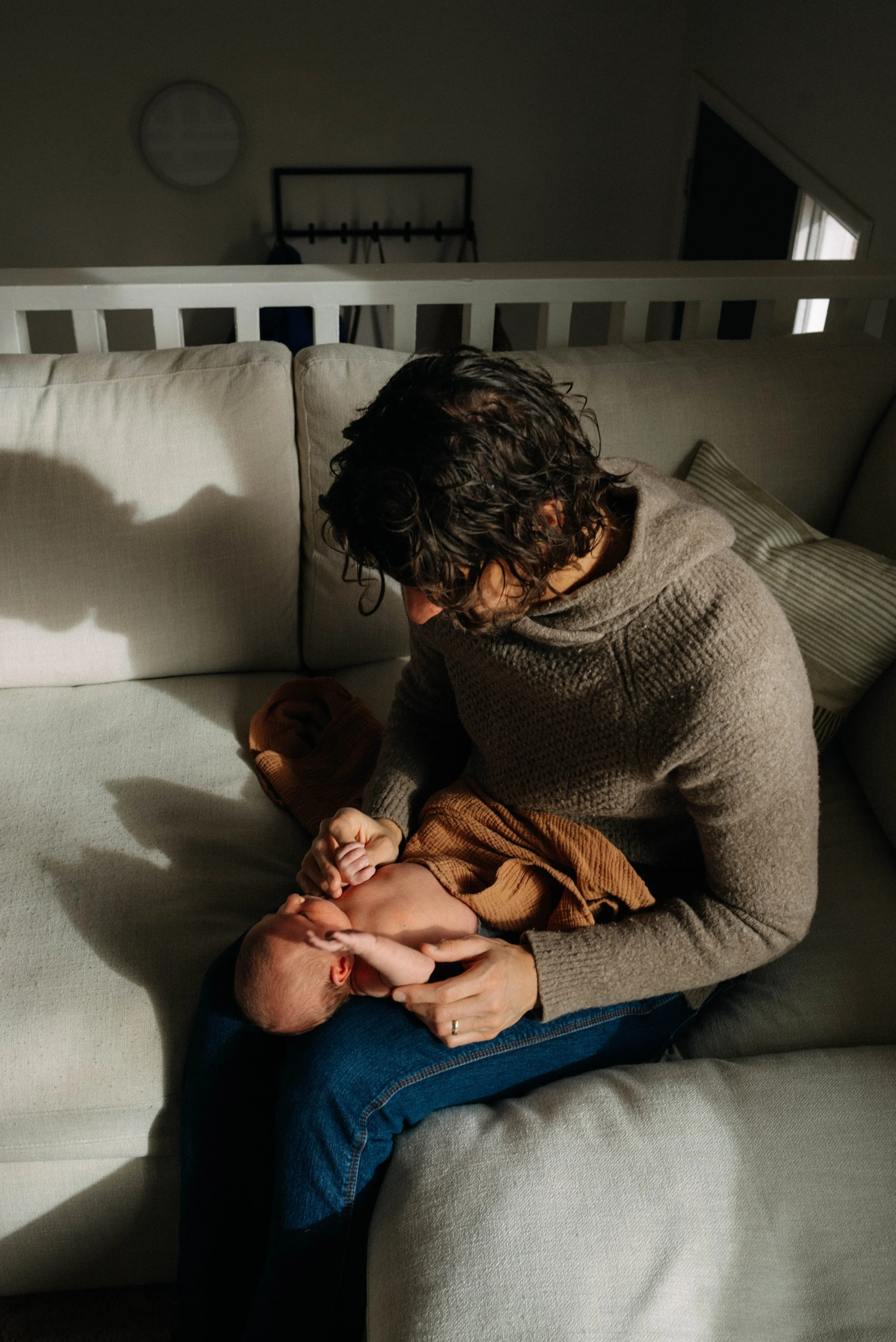 A woman sitting on a gray couch holding a baby in her lap, with sunlight coming through a window highlighting the scene.