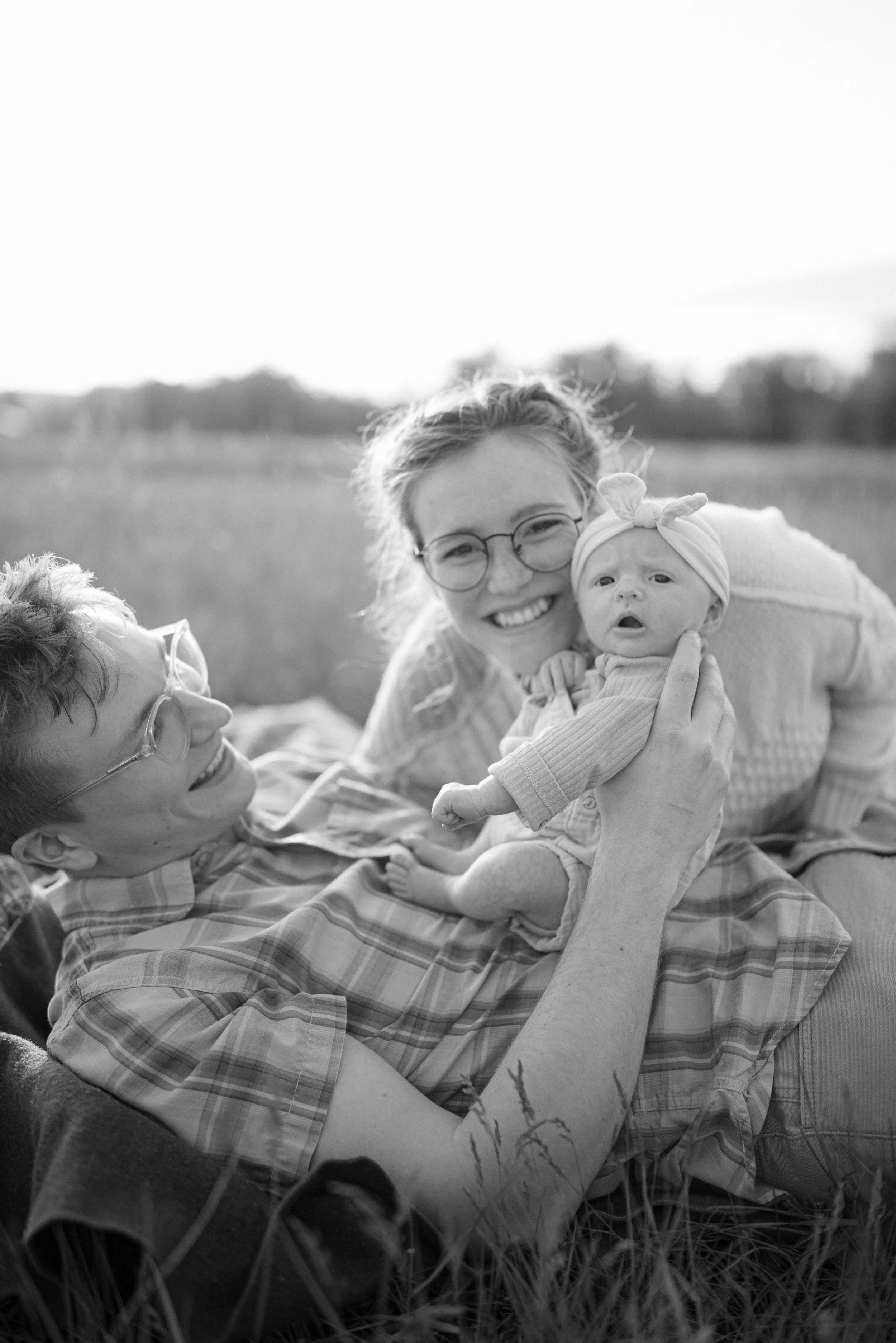 A black and white photo of mom and dad and a baby lying on the grass outdoors. Dad is holding and smiling at the baby, and mom is looking at them, all smiling. The background shows an open field in Bozeman, Montana. 