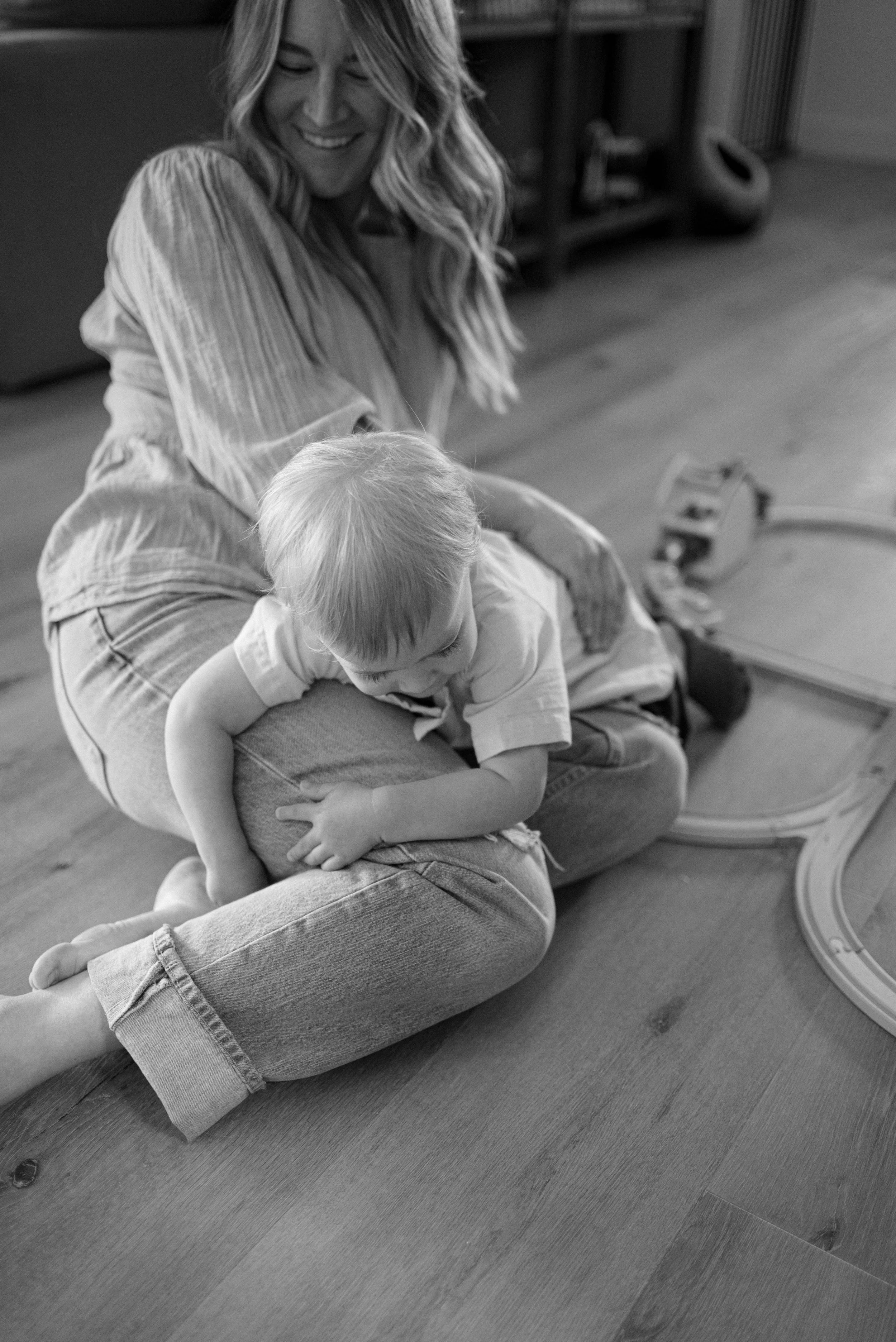 A woman sitting on a wooden floor with a young child resting on her lap. The woman is smiling and looking down at the child, who is leaning onto her with arms wrapped around her knee. There are toy train tracks visible on the floor.