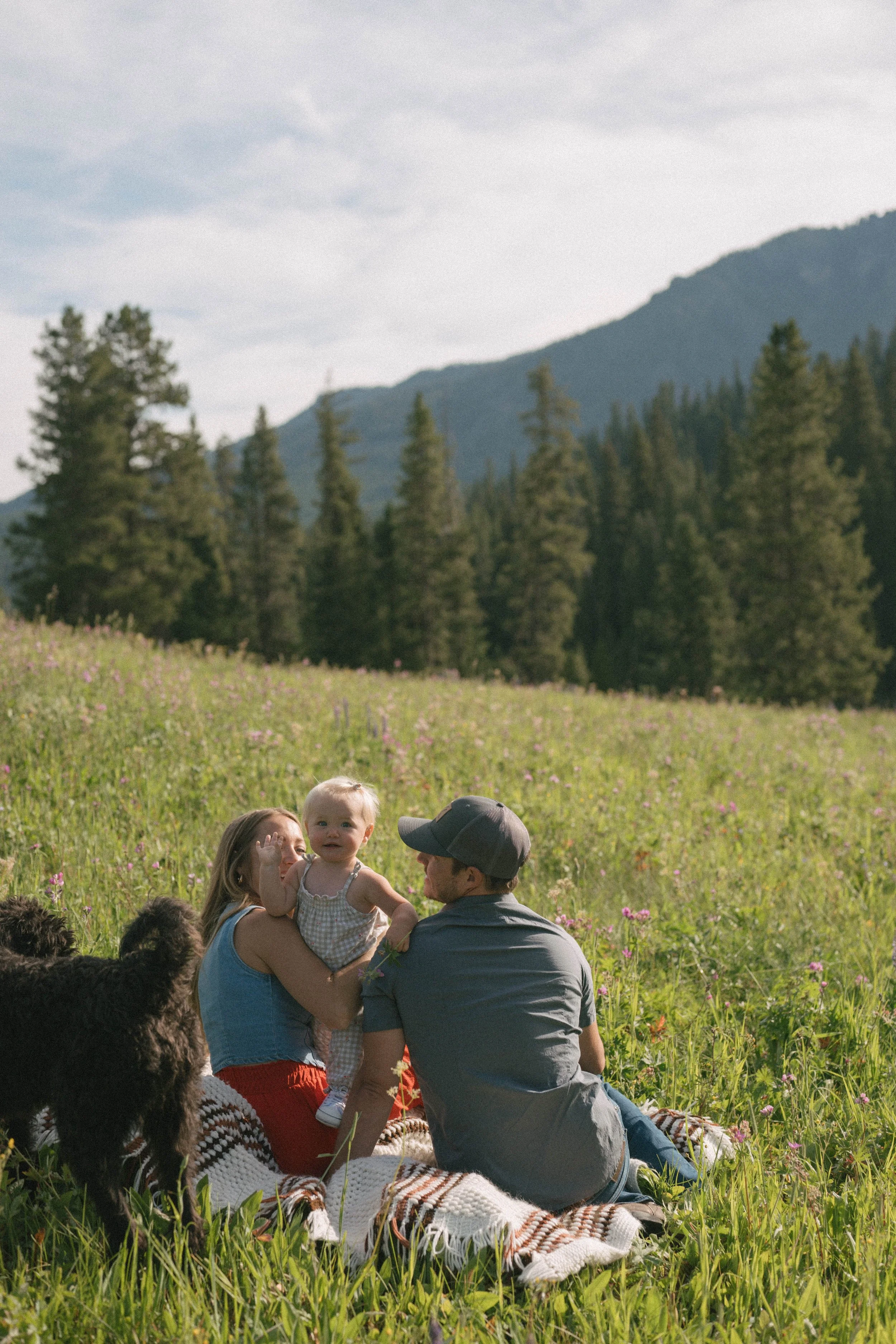 Family of two adults and a young girl sitting on a blanket in a grassy meadow with wildflowers, mountains, and trees in the background on a sunny day.