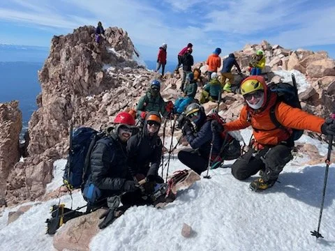 Group of climbers on a snowy mountain summit with rocky outcroppings, some taking photos and others resting, all wearing helmets and winter gear.