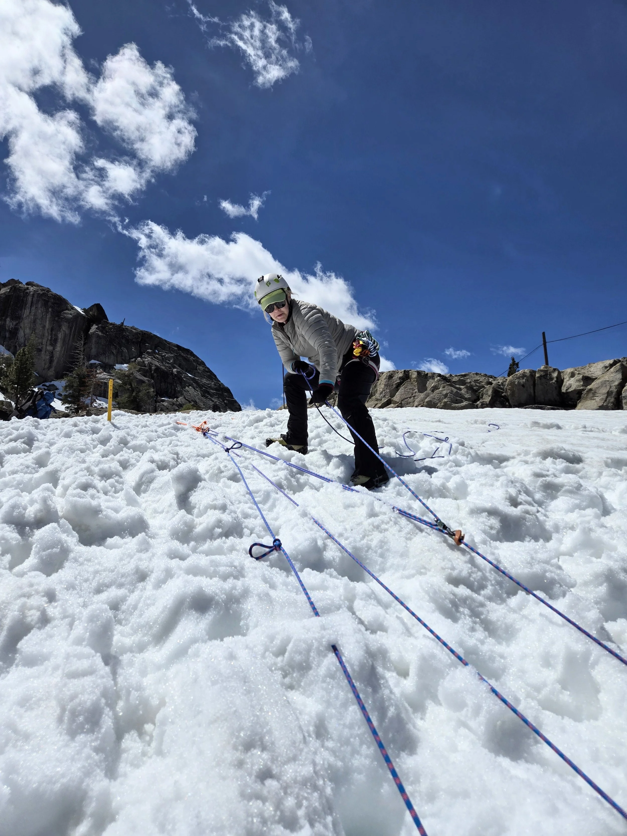 A person in winter gear, including a helmet and sunglasses, is climbing snowy mountain terrain with ropes, with rocky cliffs and blue sky with clouds in the background.