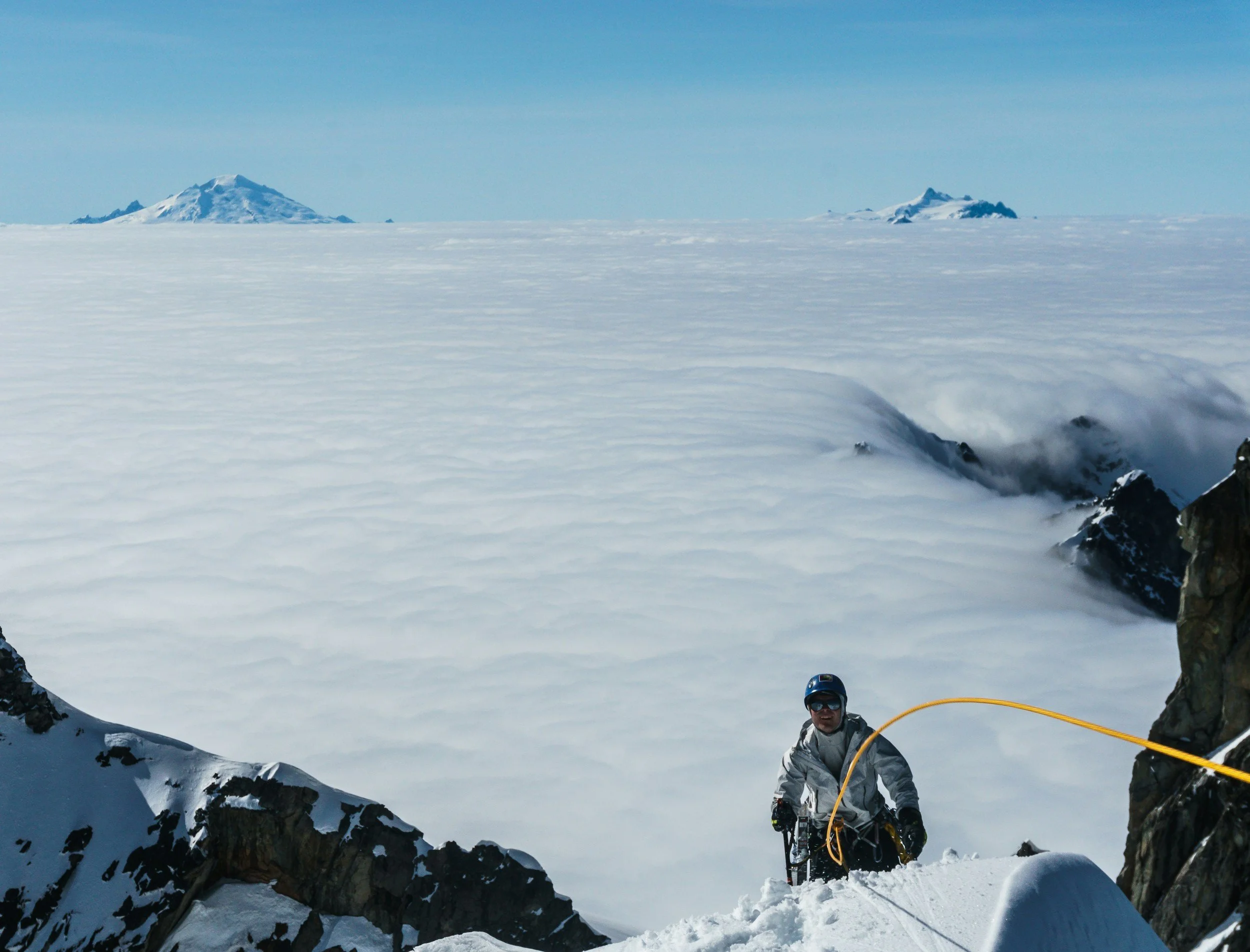 A person in winter gear climbing snowy mountain terrain, surrounded by rocks with a snow-covered mountain and a sea of clouds in the background.