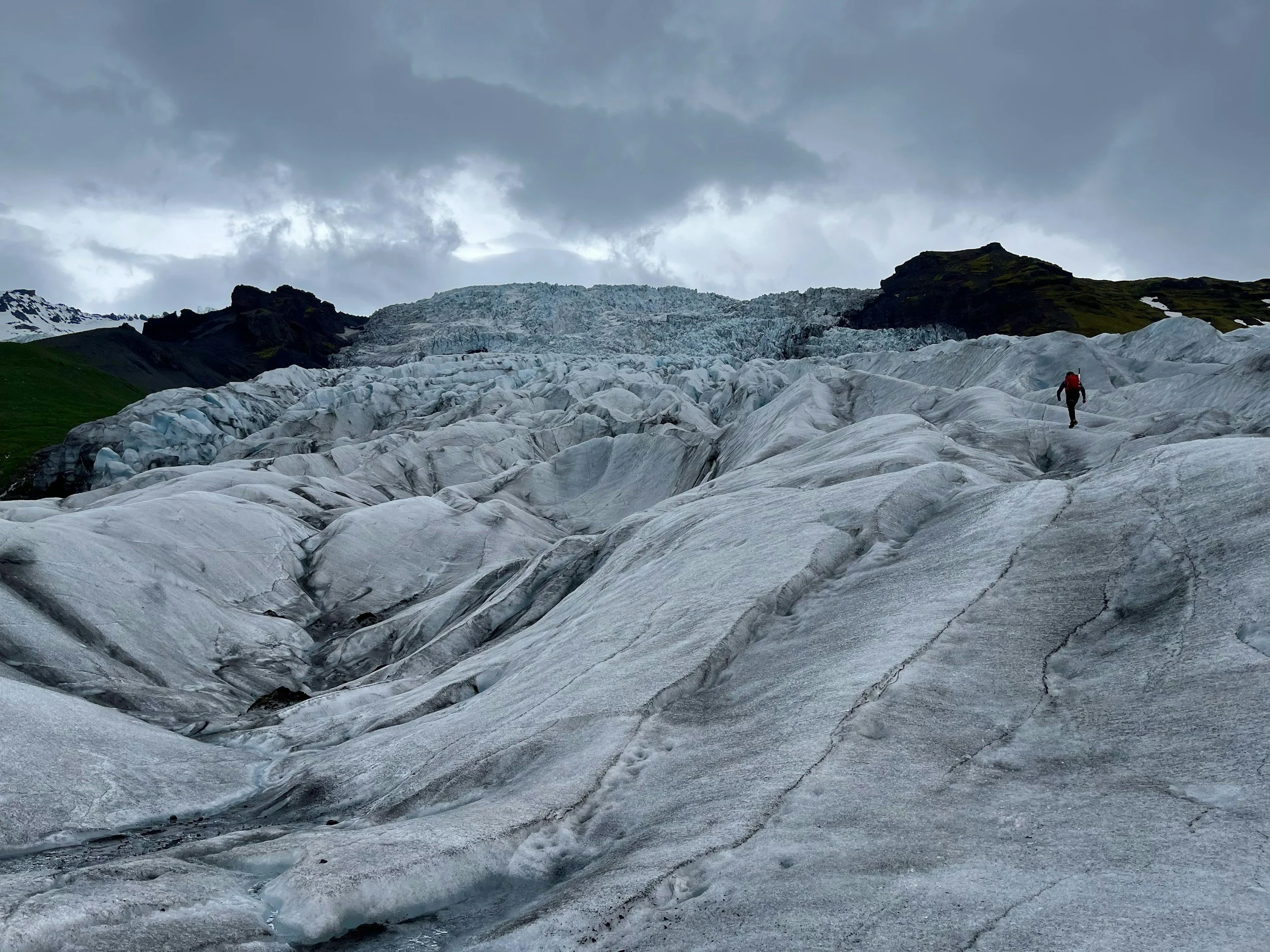 A person hiking across a large glacier under a cloudy sky, with some dark mountains in the background.