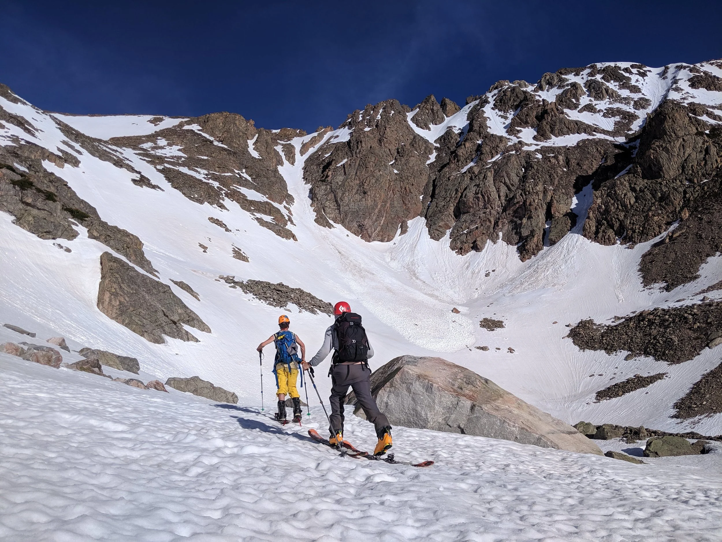 Two hikers in snow gear walking on snow with mountain rocks and snow-covered peaks in the background under a clear blue sky.