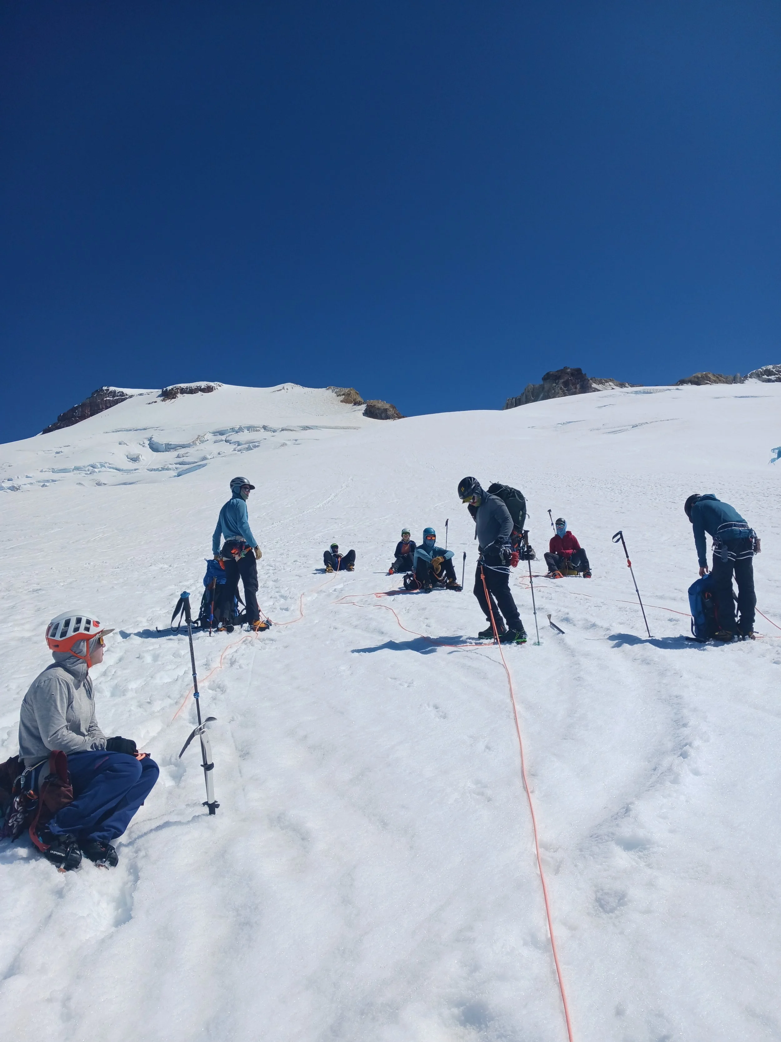Group of people preparing for a mountaineering or glacier expedition on snowy terrain with ropes, ice axes, and backpacks, under a clear blue sky.