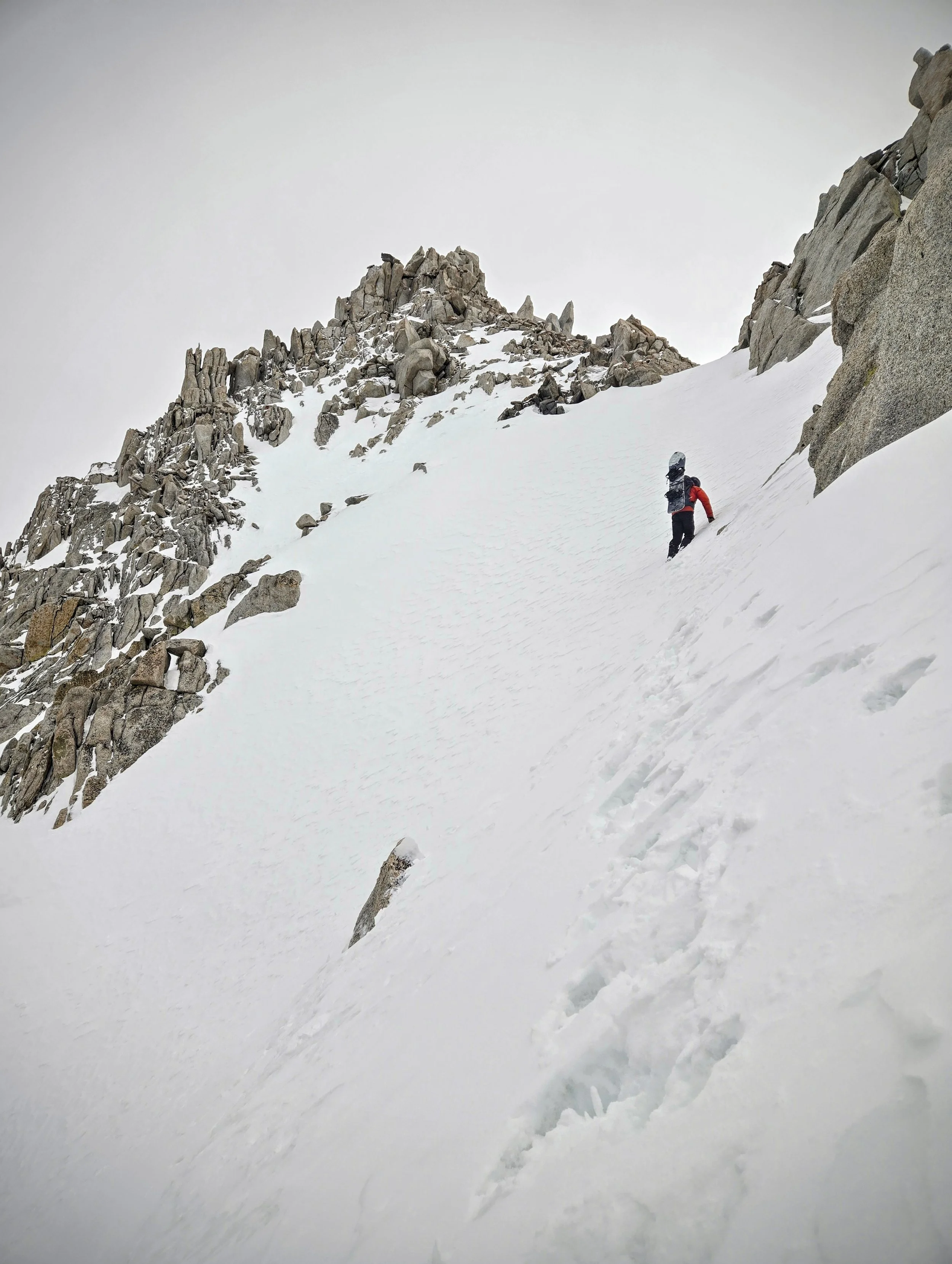 A person in outdoor gear climbing snowy mountain terrain with rocky cliffs in the background.