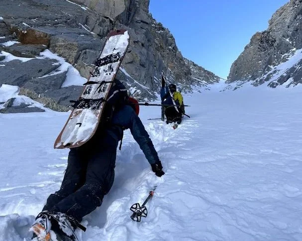 Two climbers are ascending a snow-covered mountain pass with steep rocky walls, using ice axes and crampons, carrying a snowboard on the back of the closest climber.