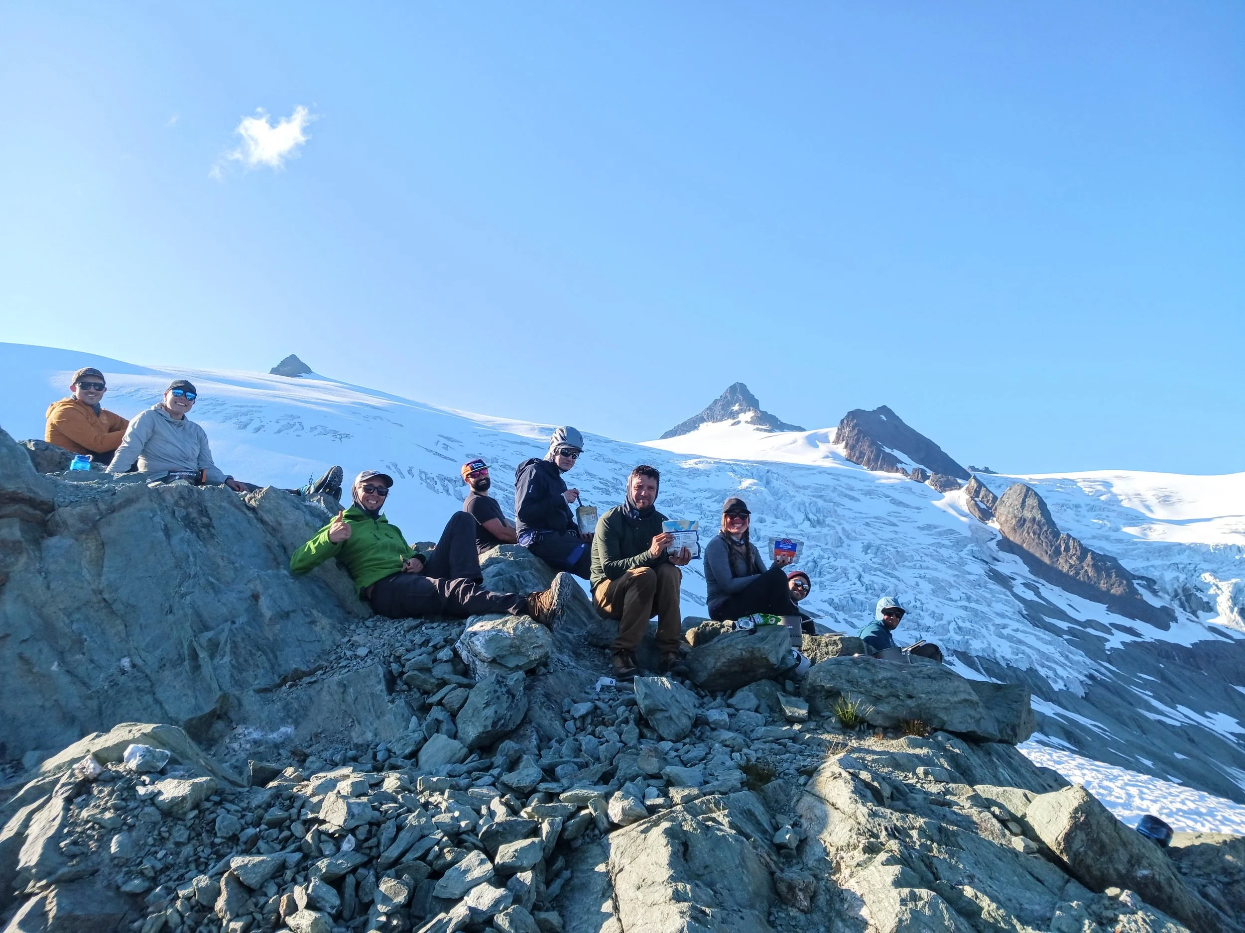 A group of ten people sitting on rocks on a snowy mountain with peaks in the background, enjoying the view and holding snacks or drinks.