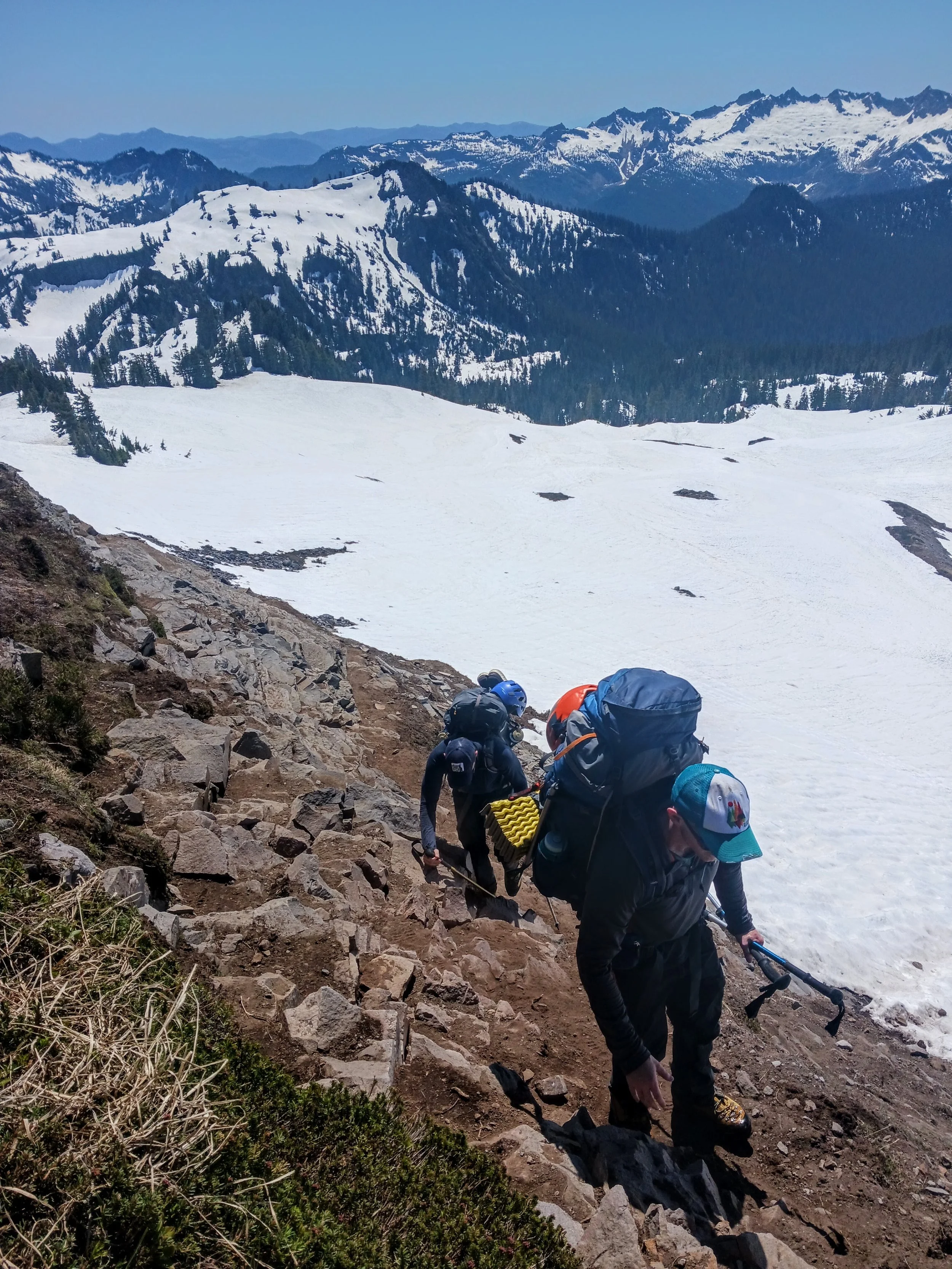 Group of hikers climbing a rocky mountain trail above snow-covered terrain with mountainous landscape in the background.