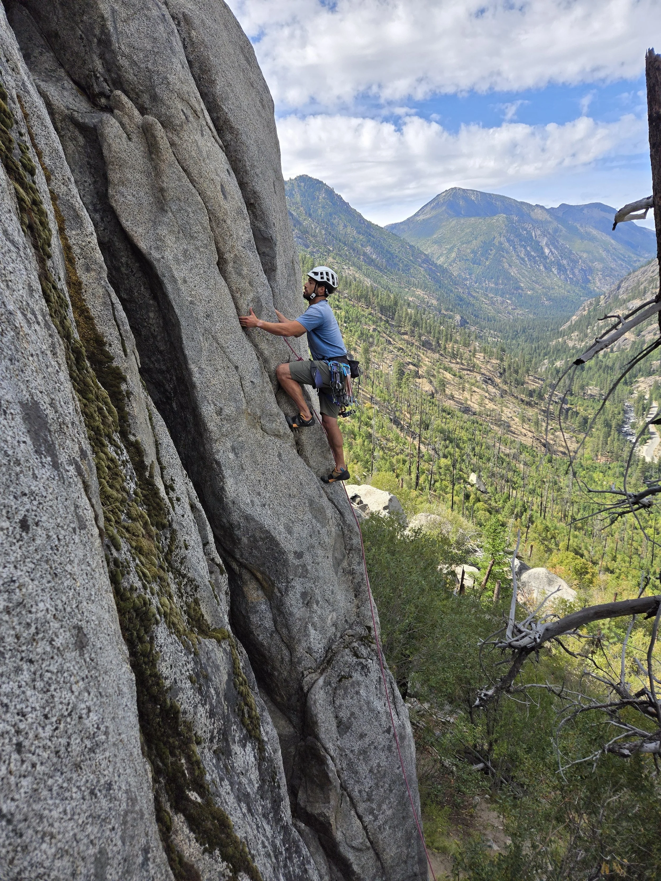 A man wearing a helmet rock climbing on a steep granite cliffside in a mountainous landscape with trees and distant peaks under a partly cloudy sky.